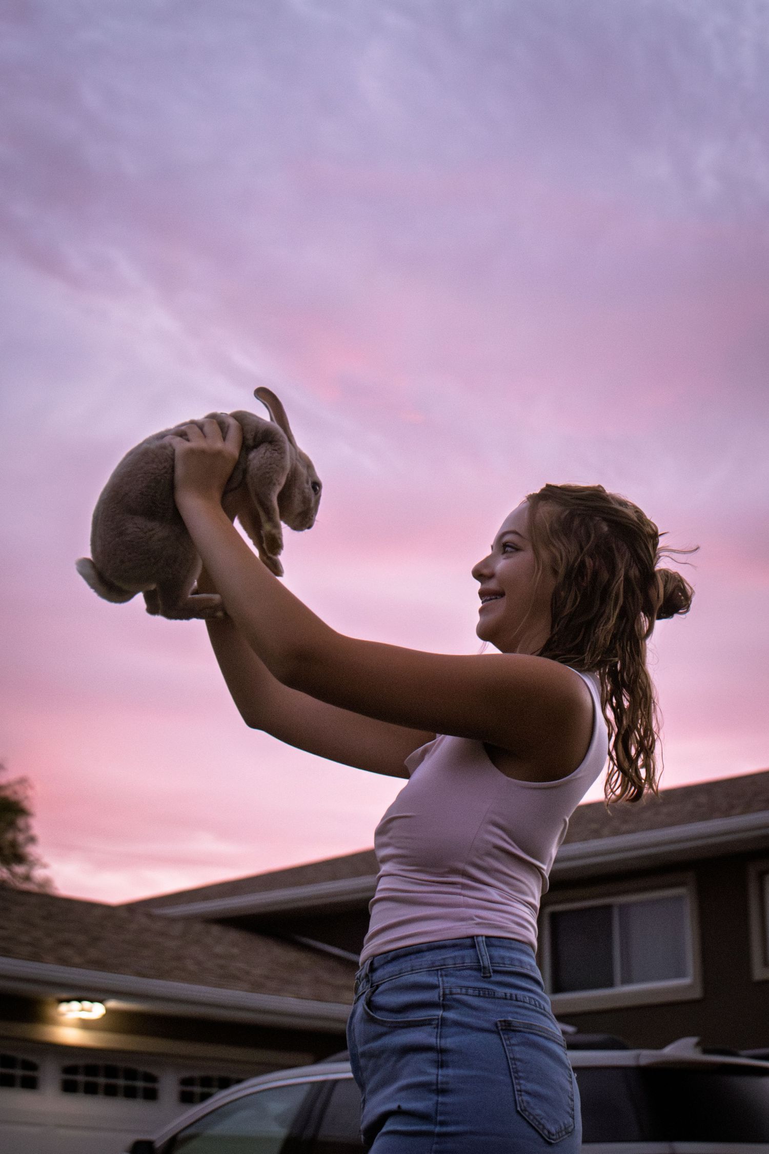 Girl holding a rabbit above her head against a pink sunset sky in a suburban neighborhood.