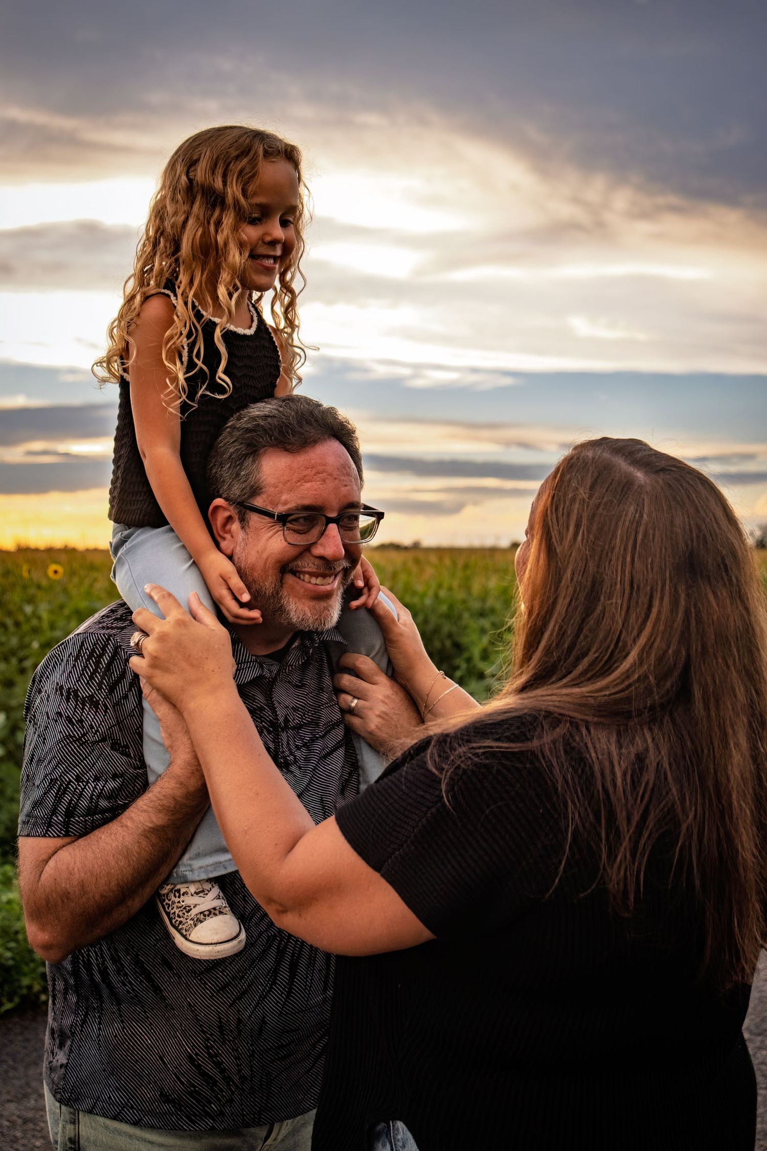 Man with glasses smiles as young girl sits on his shoulders at sunset while woman reaches up to them.
