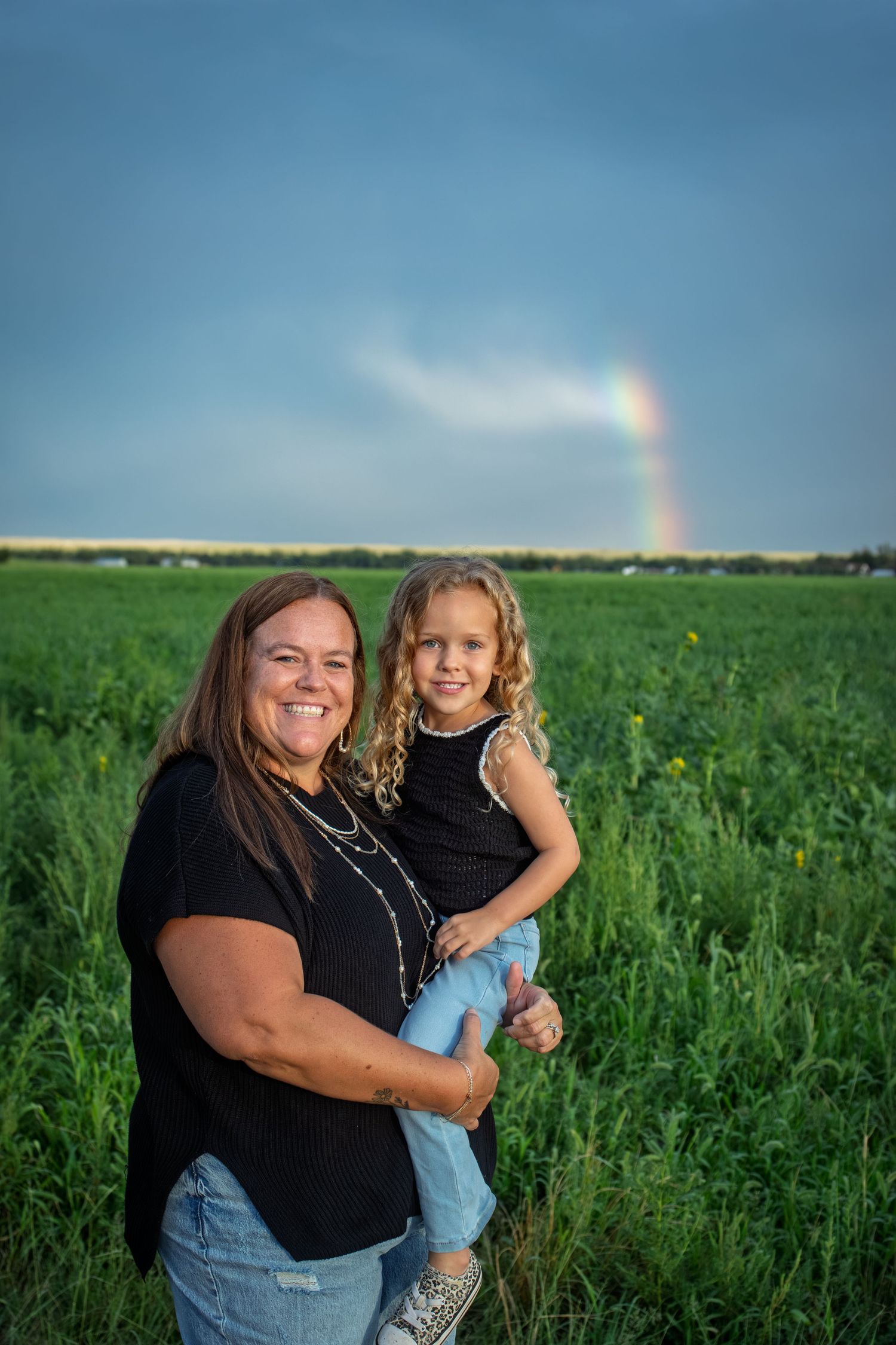 Smiling woman in black shirt holding young curly-haired girl in green field with rainbow in sky.