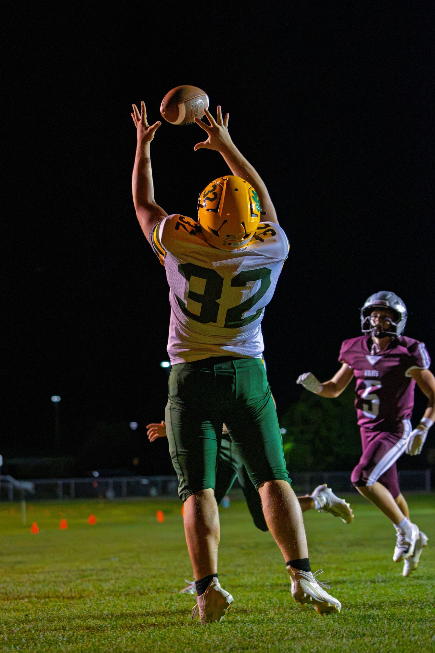 Football player #32 in green and gold uniform leaps to catch a pass during a night game.