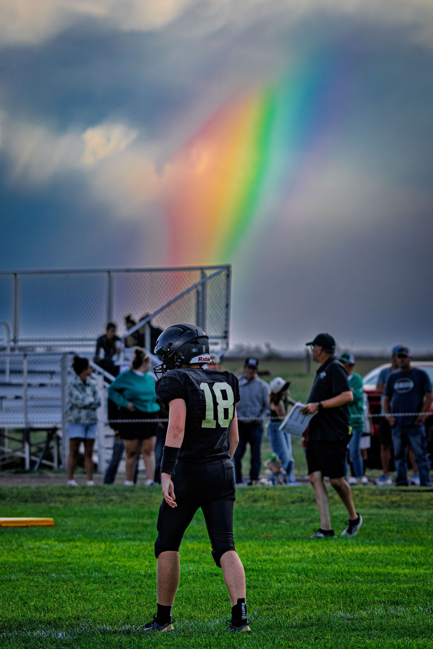 Football player #18 in black uniform stands on green field with vibrant rainbow in stormy sky behind bleachers.