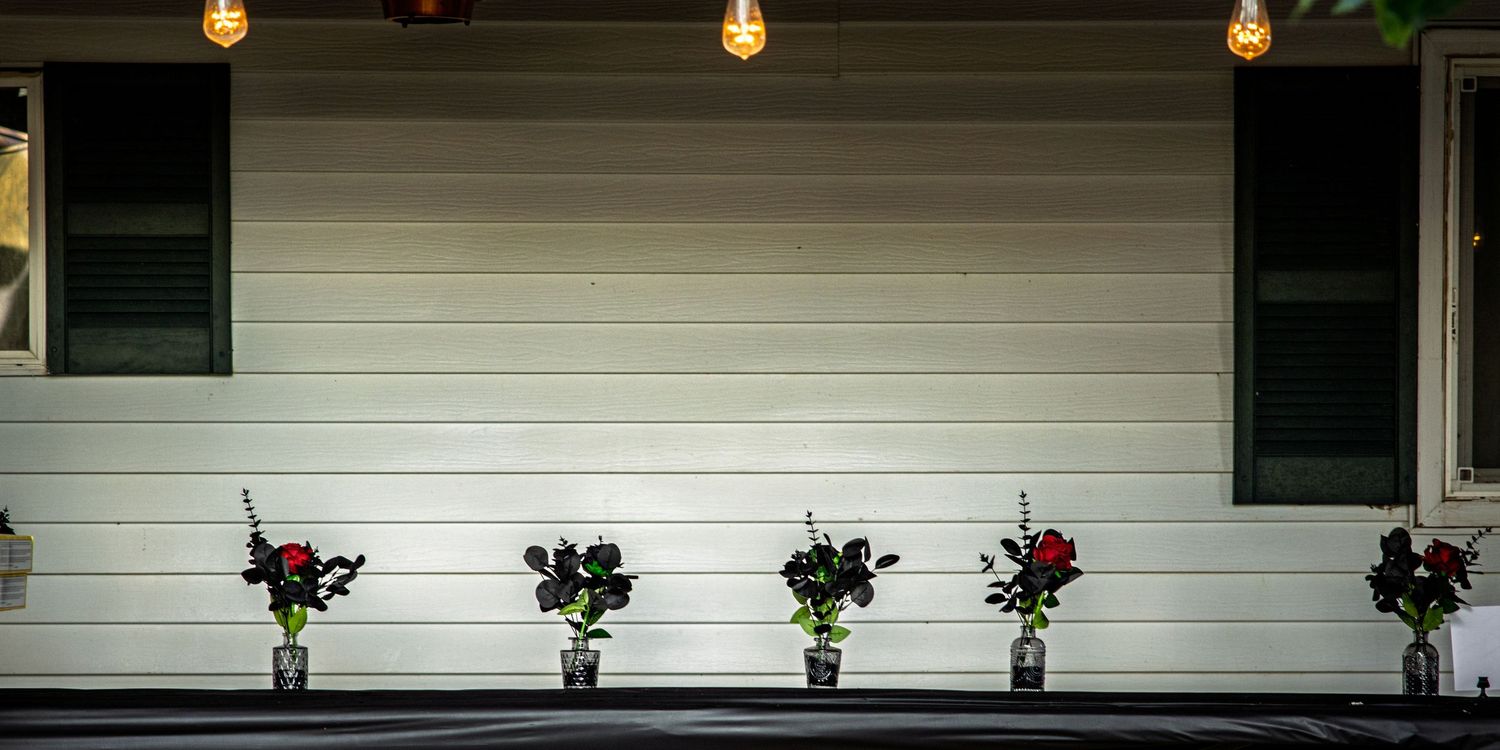 Row of flower arrangements in glass vases lined up against a white siding wall under warm string lights at night.
