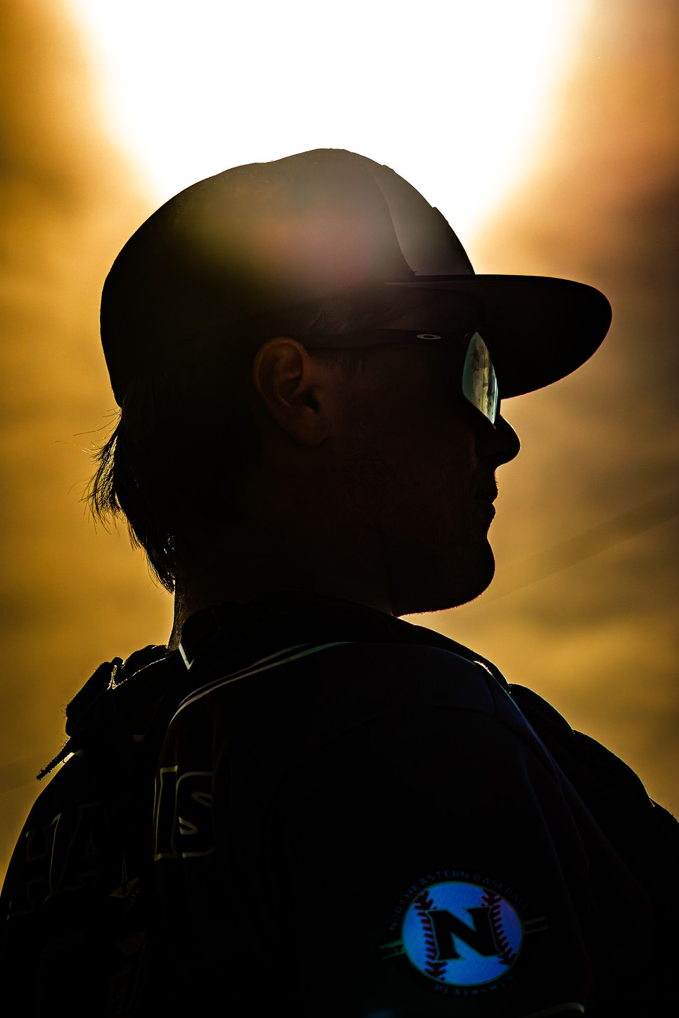Dramatic silhouette of a baseball player wearing sunglasses and cap against a golden sunset sky.