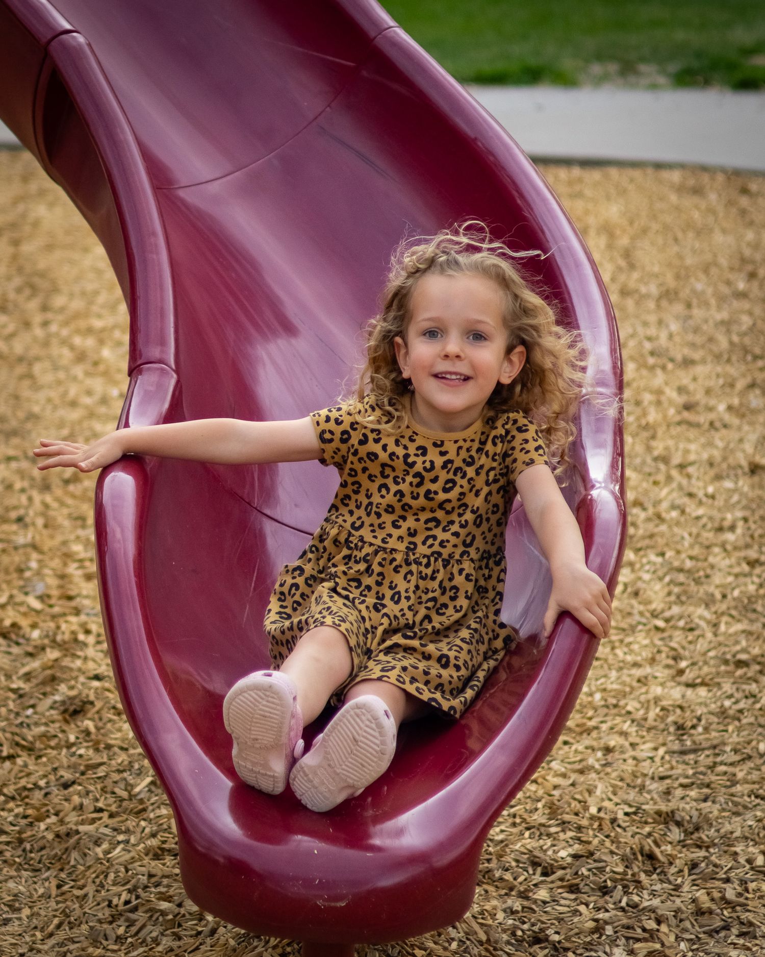 A happy young girl with curly hair sliding down a red playground slide in a leopard print dress.