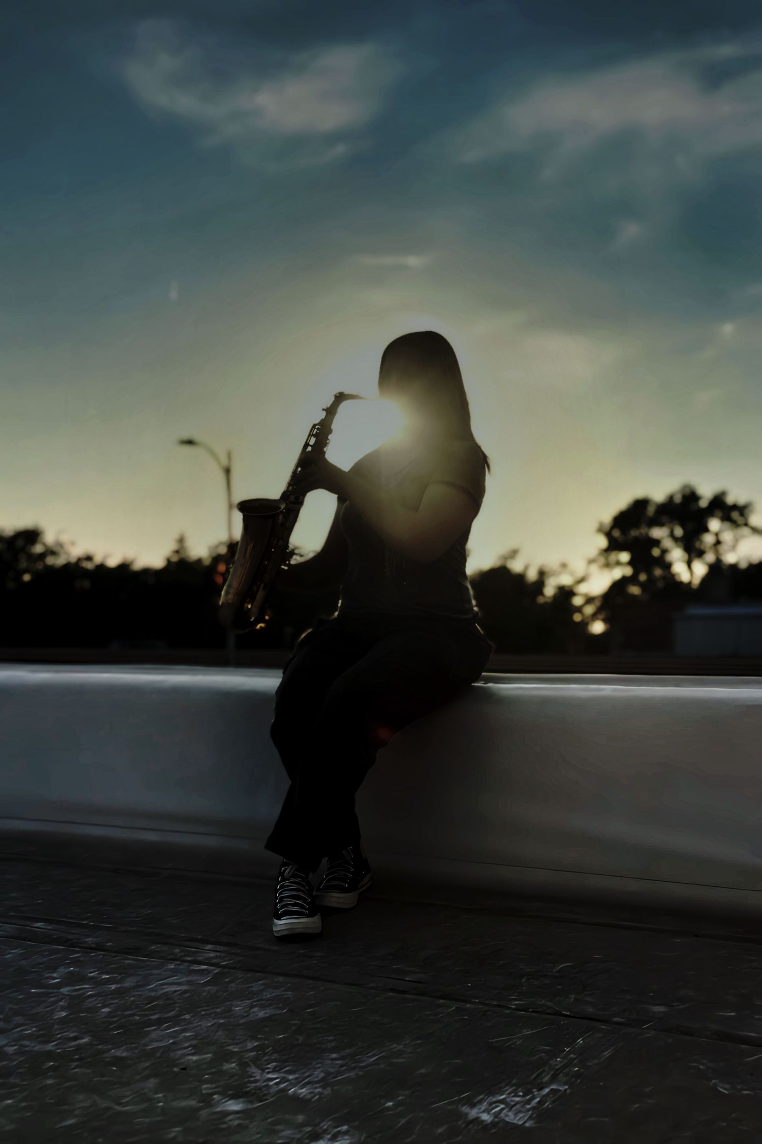 Silhouette of a saxophonist sitting on a ledge playing at sunset with dramatic backlit golden sky.