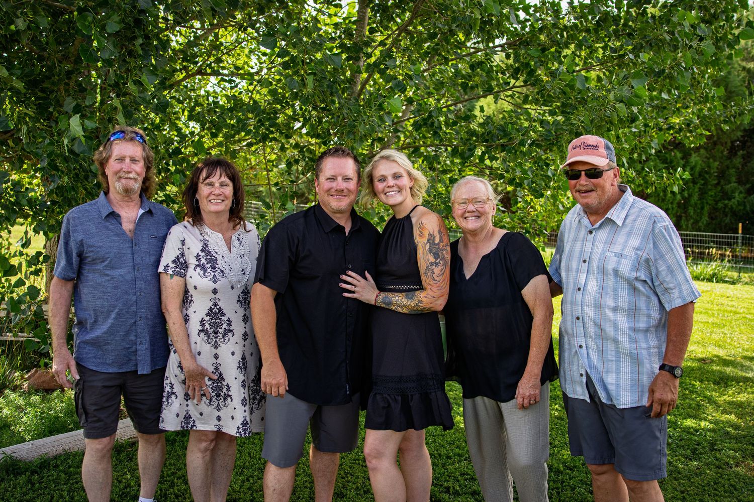 Six family members smiling together outdoors in a garden with lush green trees behind them.