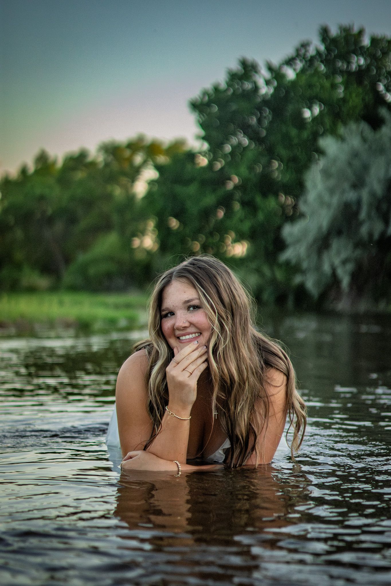 Smiling young woman lying in shallow river water surrounded by lush green trees at dusk.