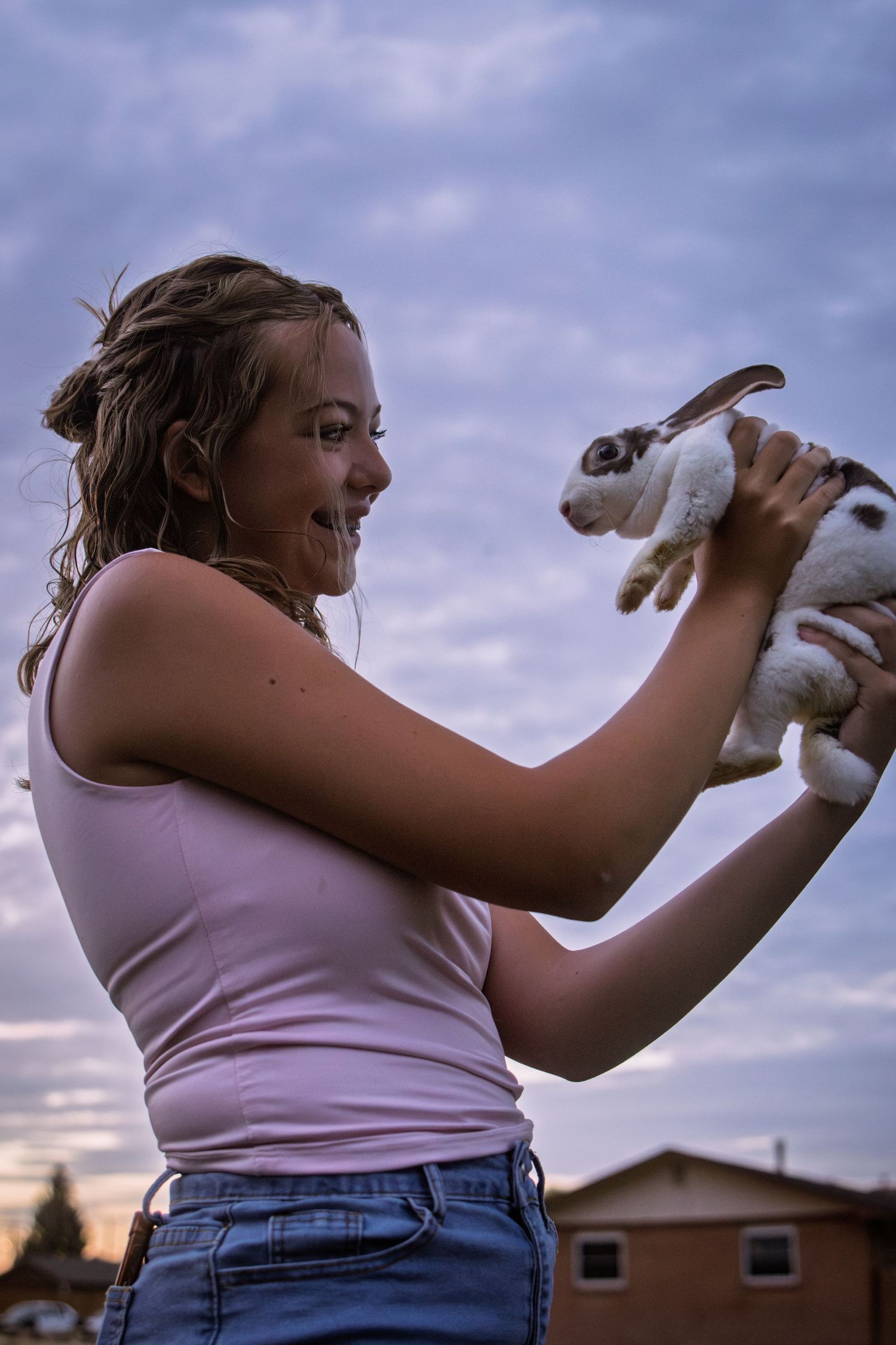 Smiling girl in pink tank top holding a white and brown rabbit up at dusk.