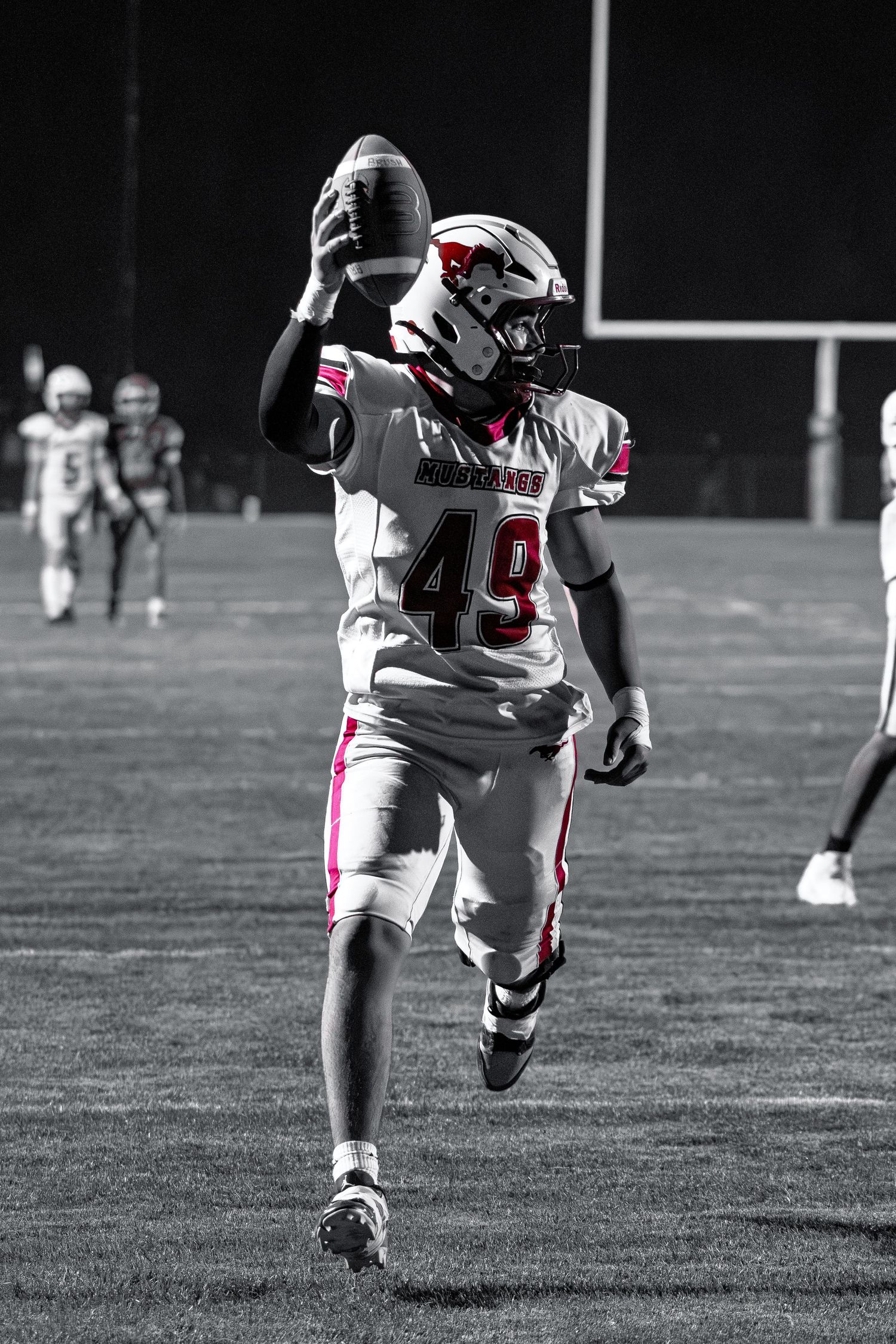 Selective color photo of football player #49 in white uniform holding ball aloft on dark field at night.