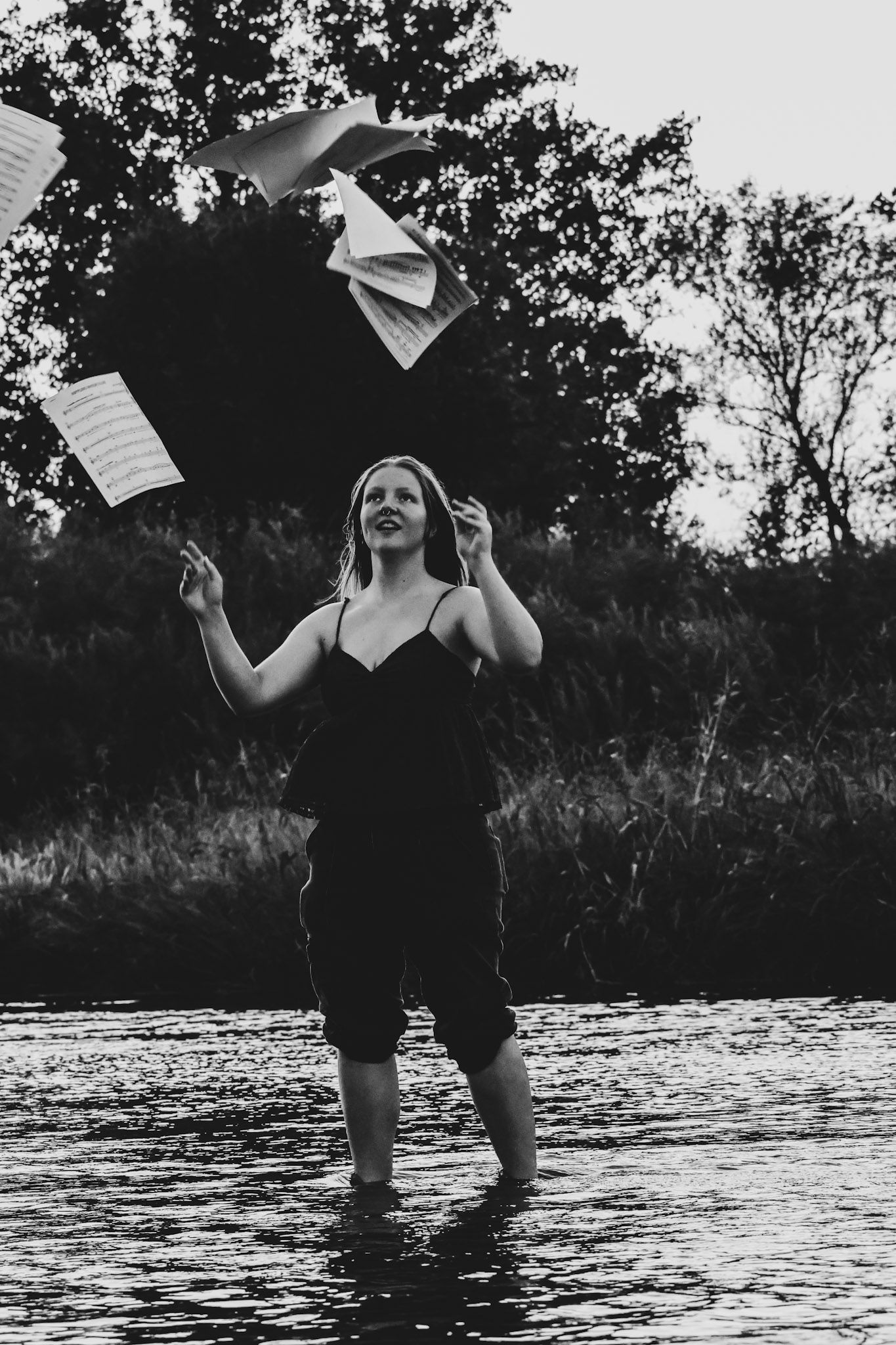 Black and white photo of a woman standing in a river tossing sheet music into the air.