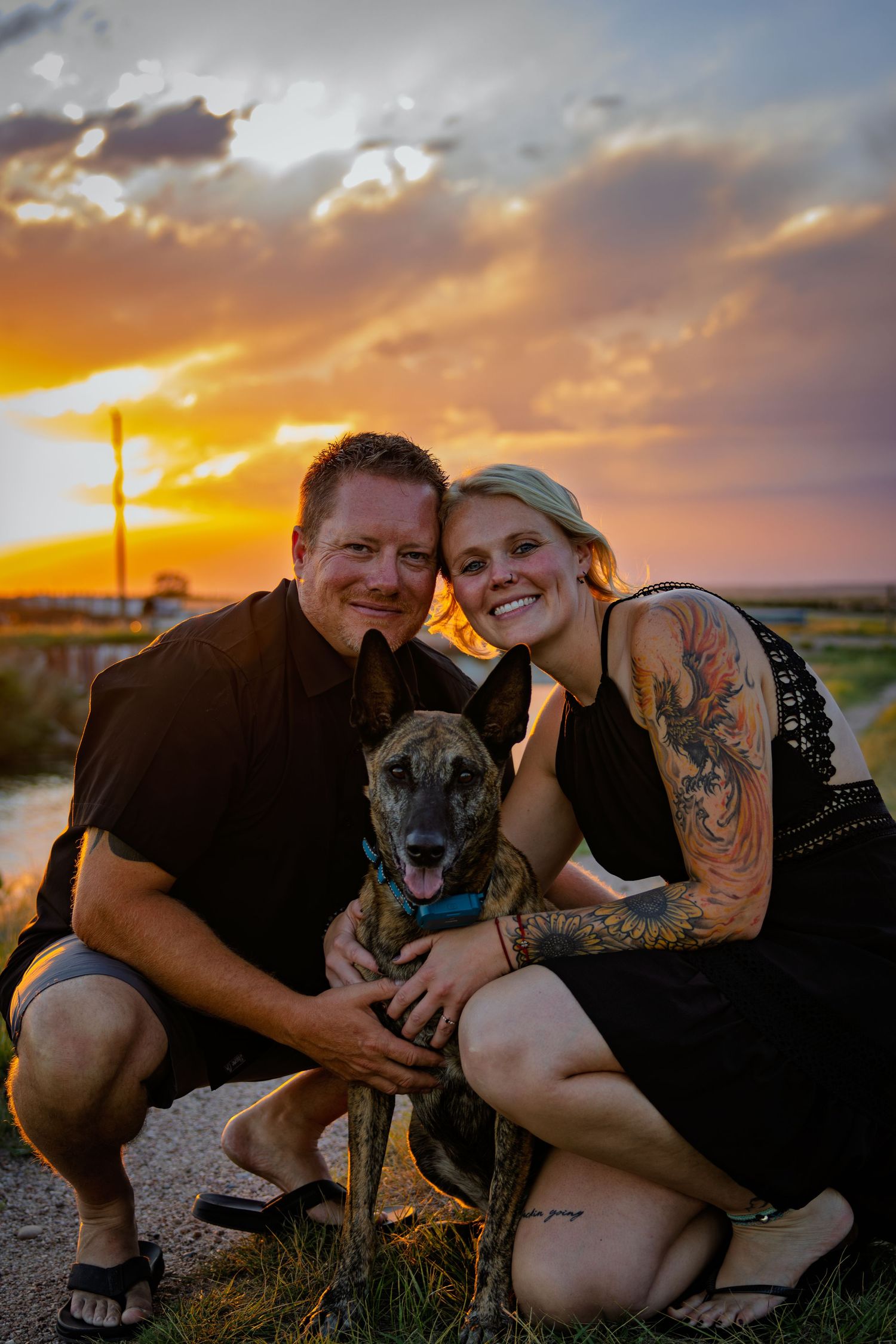 Smiling couple crouching with their brindle dog during a stunning golden sunset outdoors.