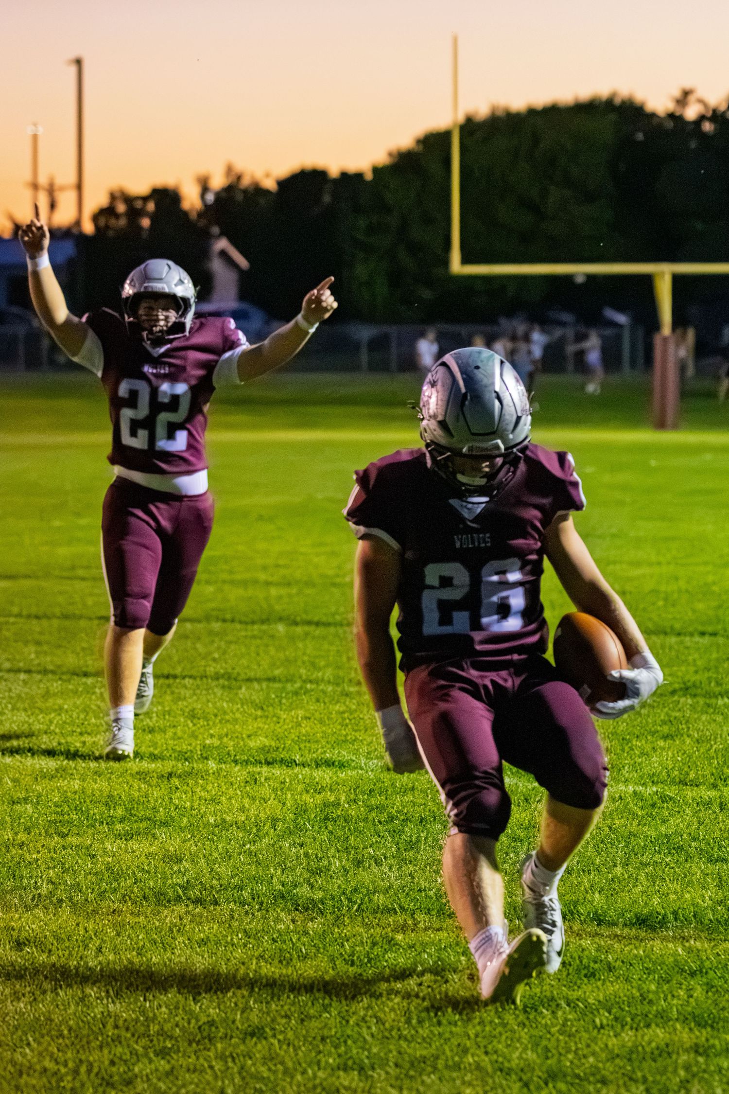 Two maroon-uniformed football players celebrate on the field at sunset, with player #22 raising arms in victory.
