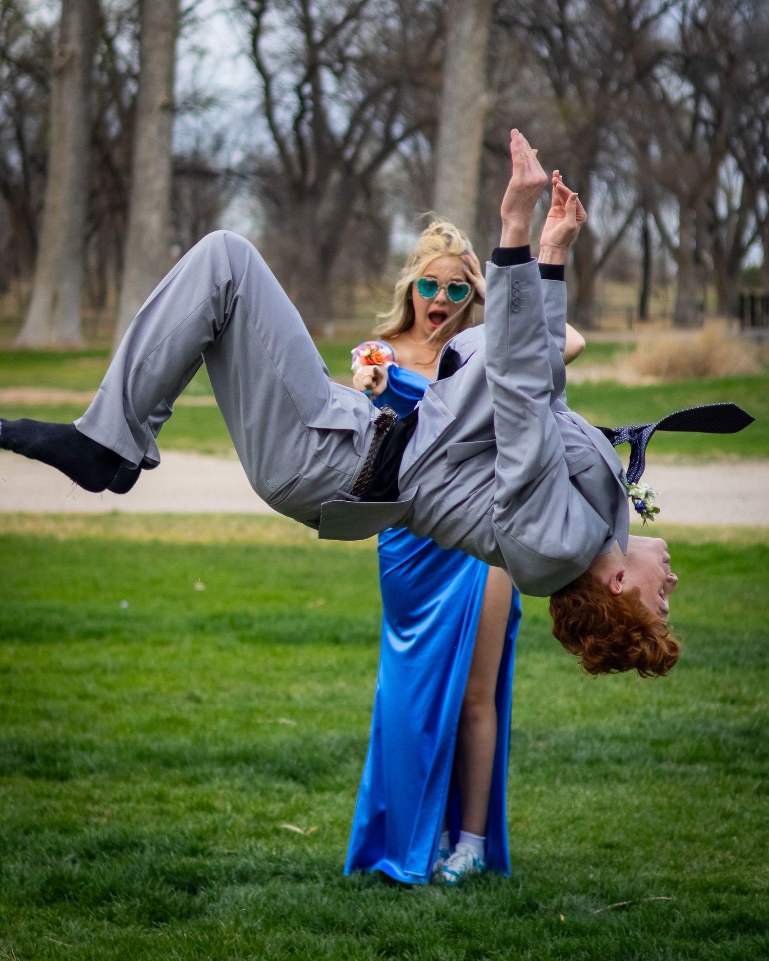 Prom couple posing playfully outdoors as boy in gray suit performs acrobatic stunt for girl in blue gown.