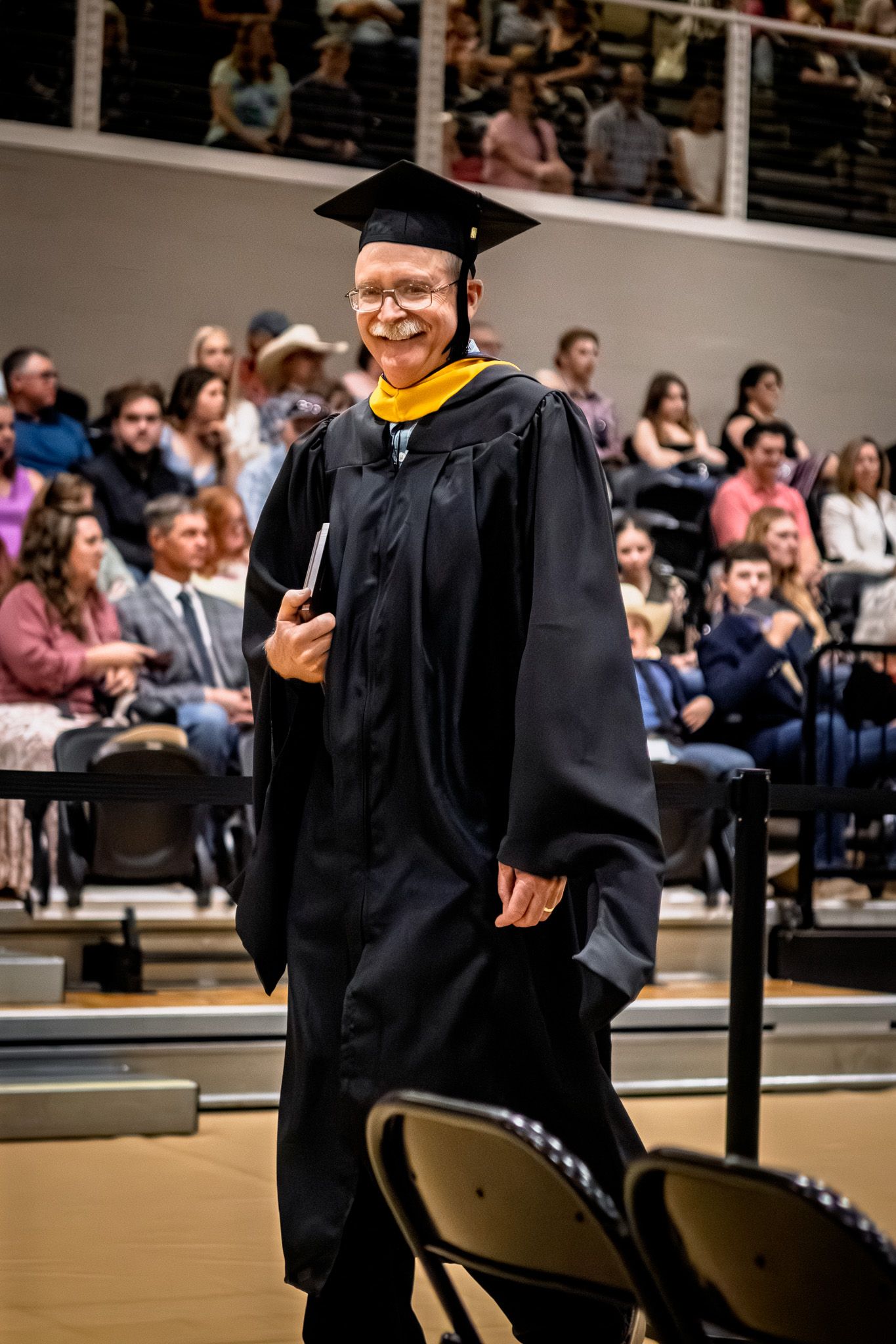 Smiling graduate in black cap and gown with yellow hood walking at commencement ceremony.