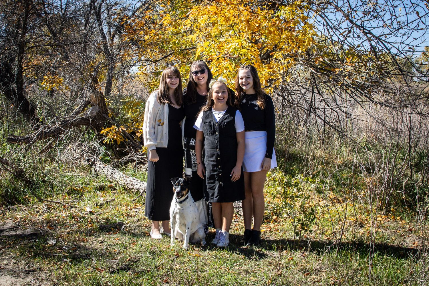 A family of three women and a girl pose with their dog under golden autumn foliage outdoors.