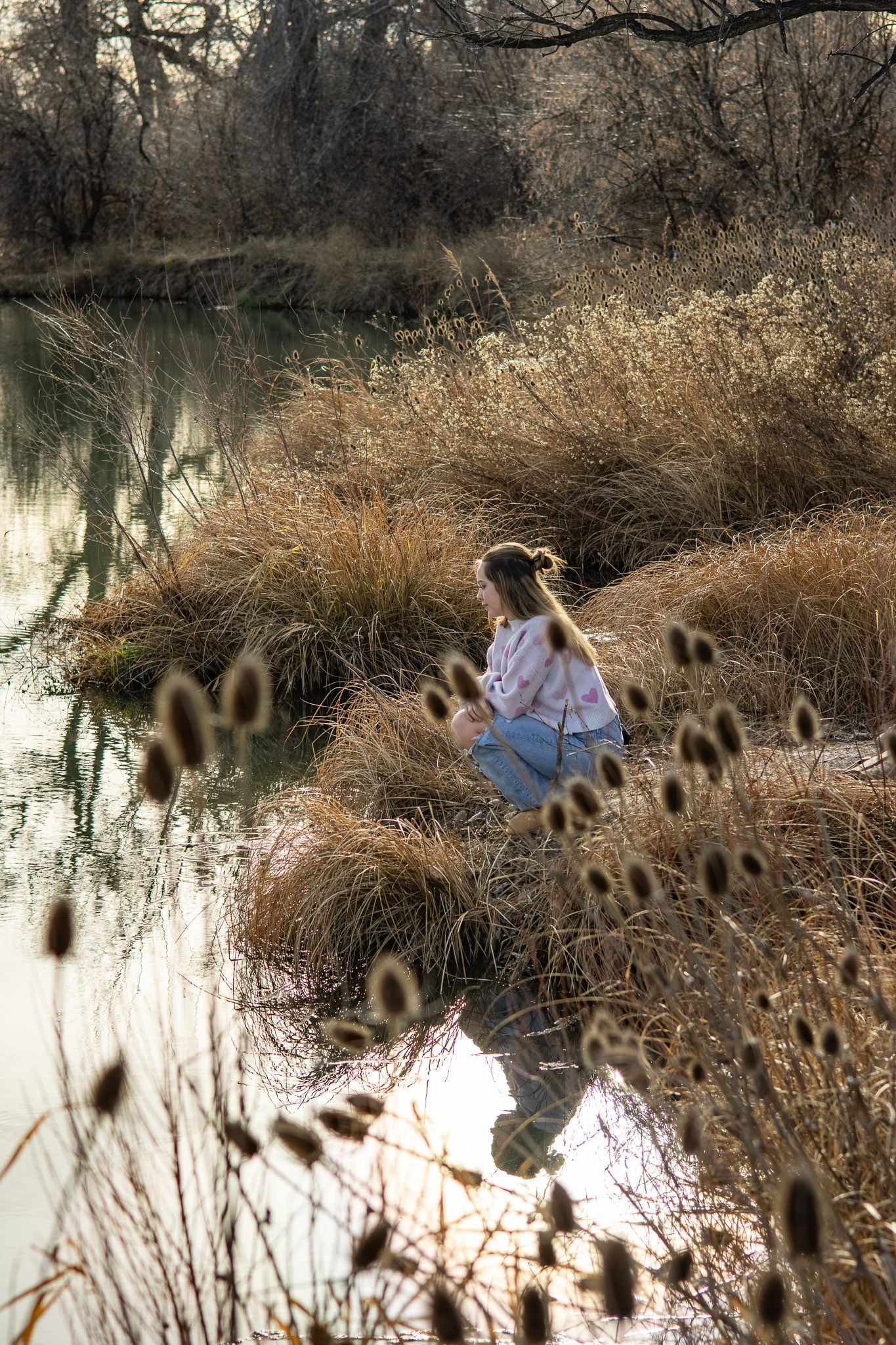A young girl crouches by a tranquil autumn river surrounded by golden dry grasses and bare trees.