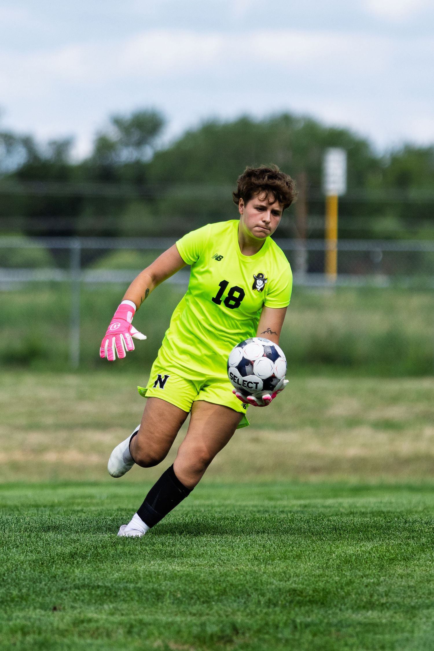 Youth goalkeeper in neon yellow jersey number 18 catching soccer ball during outdoor game.