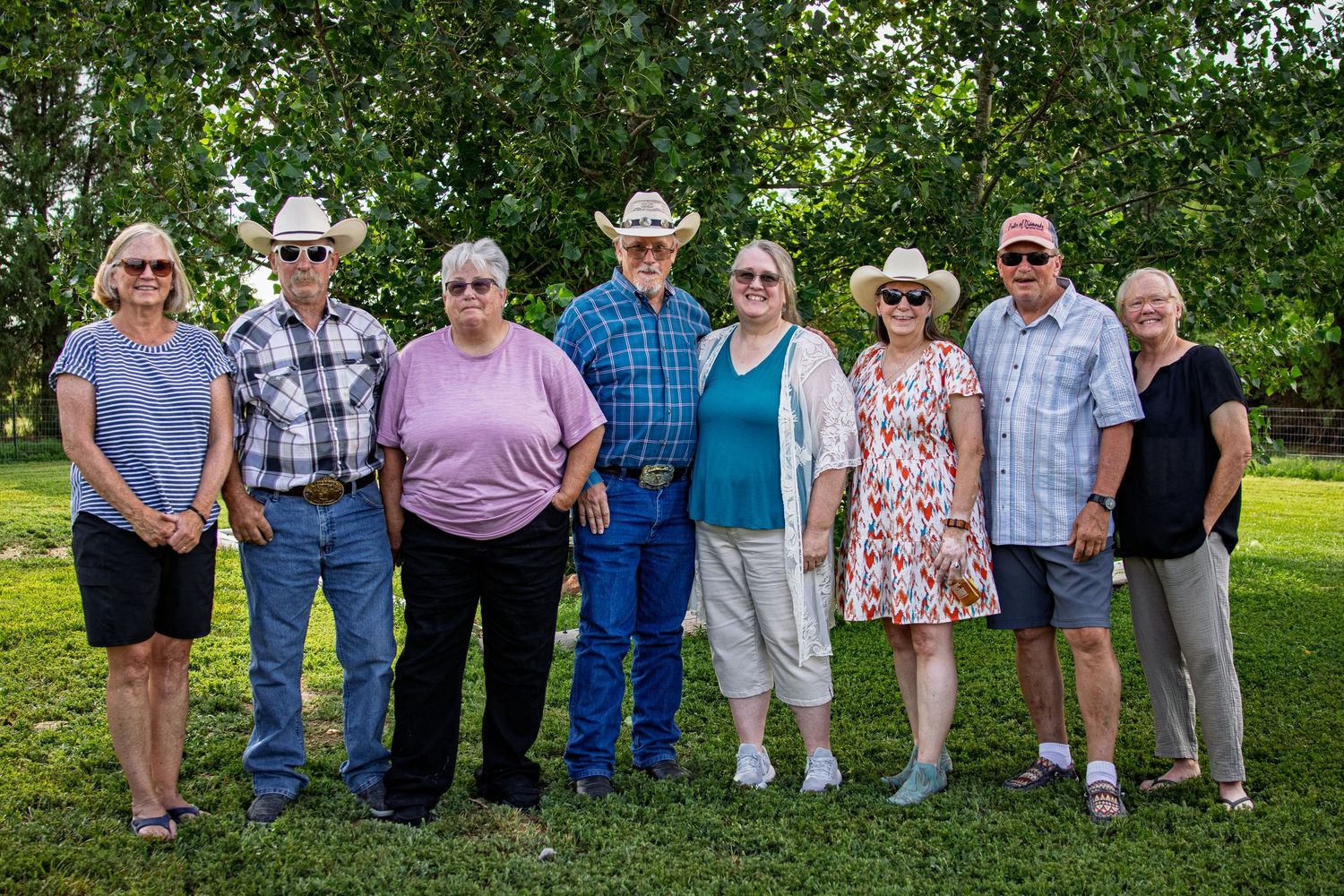Eight older adults wearing hats posing together outdoors on green grass with trees behind.