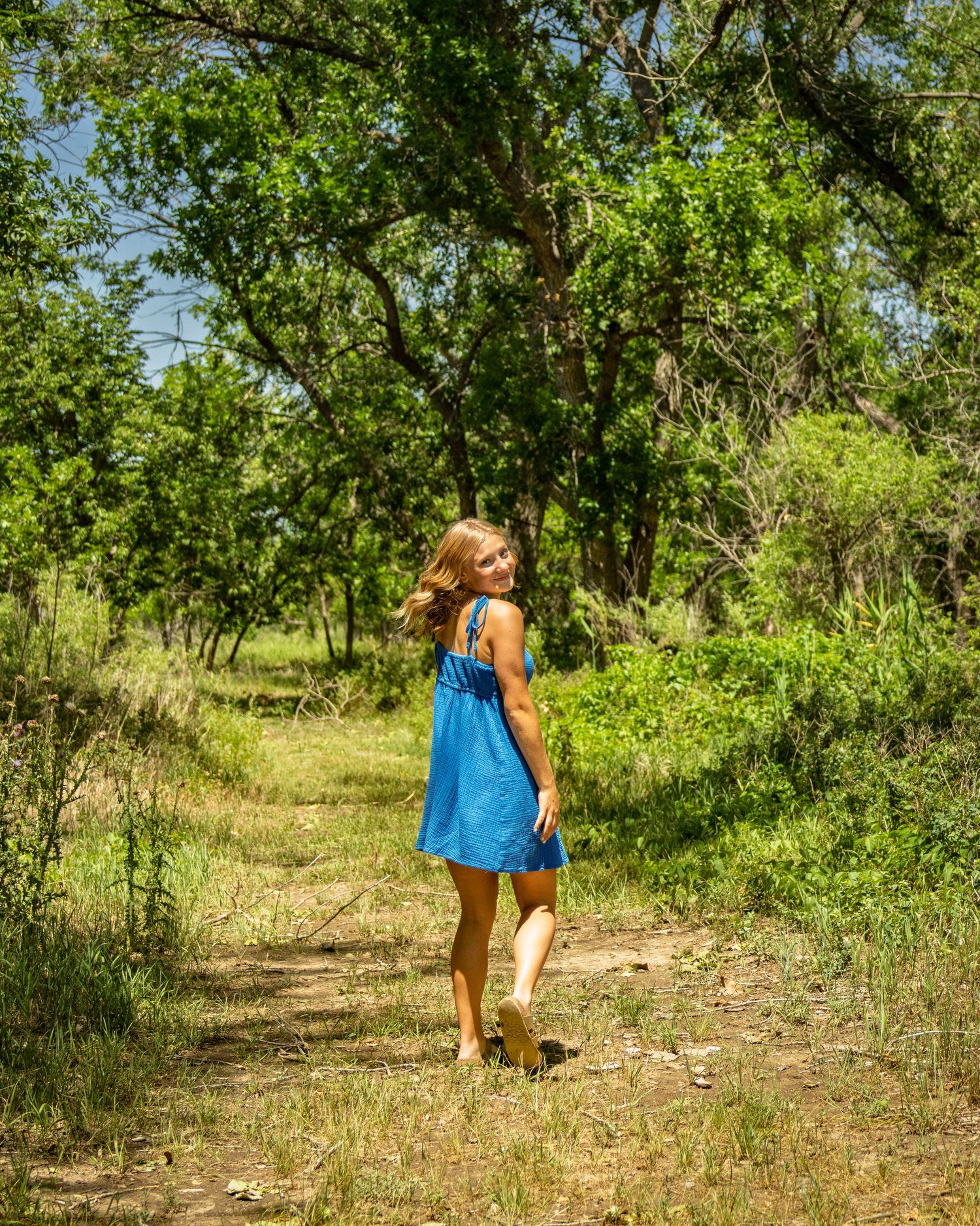Young woman in blue dress walking on a sunny wooded trail surrounded by green trees.