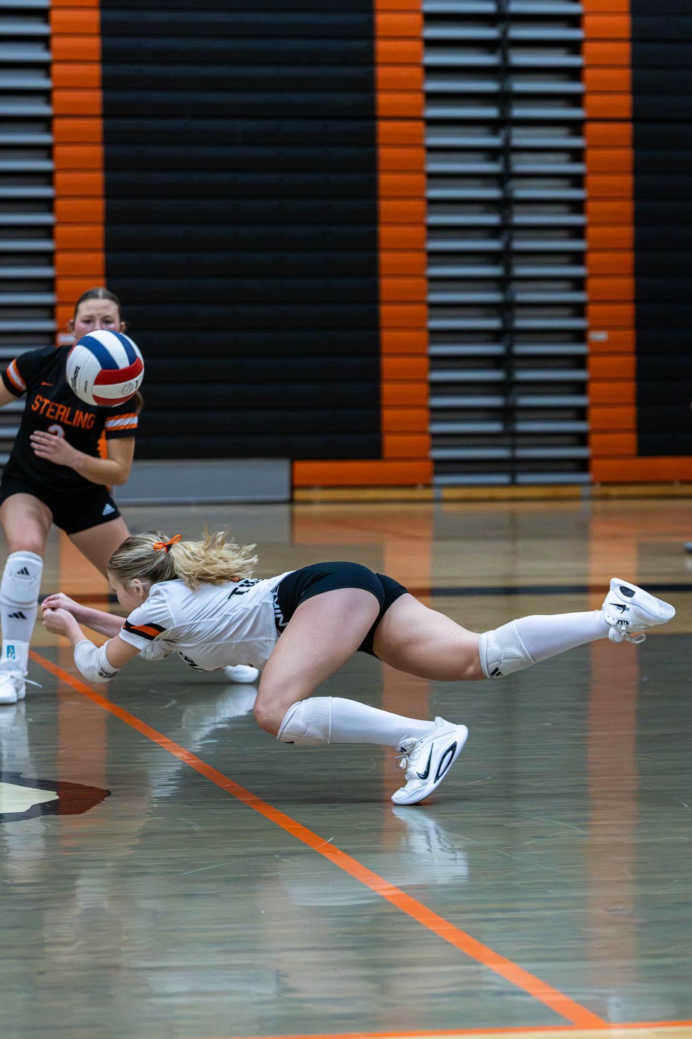 Female volleyball player dives to the floor to dig the ball during a competitive match.