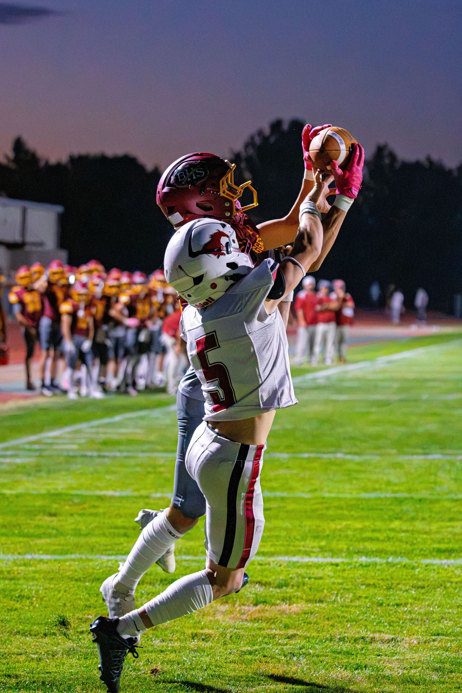 Football player in red and white uniform leaps to make a catch during a night game under purple sky.