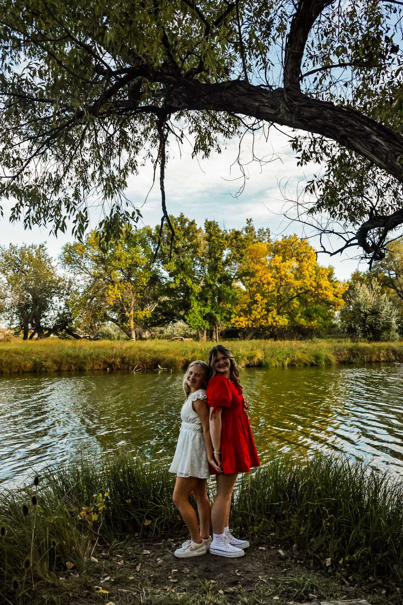 Two girls in white and red dresses stand holding hands by a scenic autumn pond under a tree.