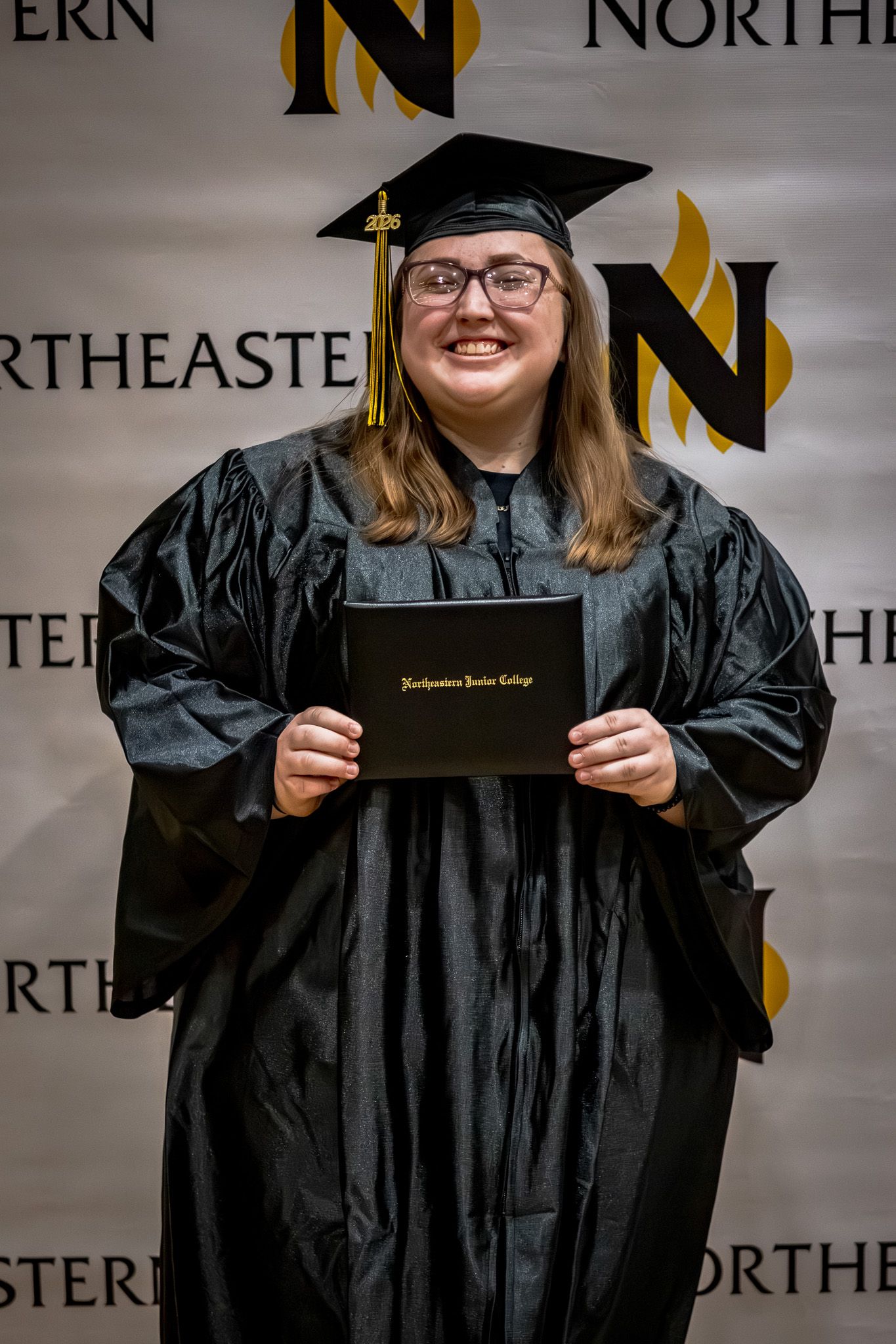 Proud female graduate wearing glasses holds Northeastern Junior College diploma in black cap and gown at ceremony.