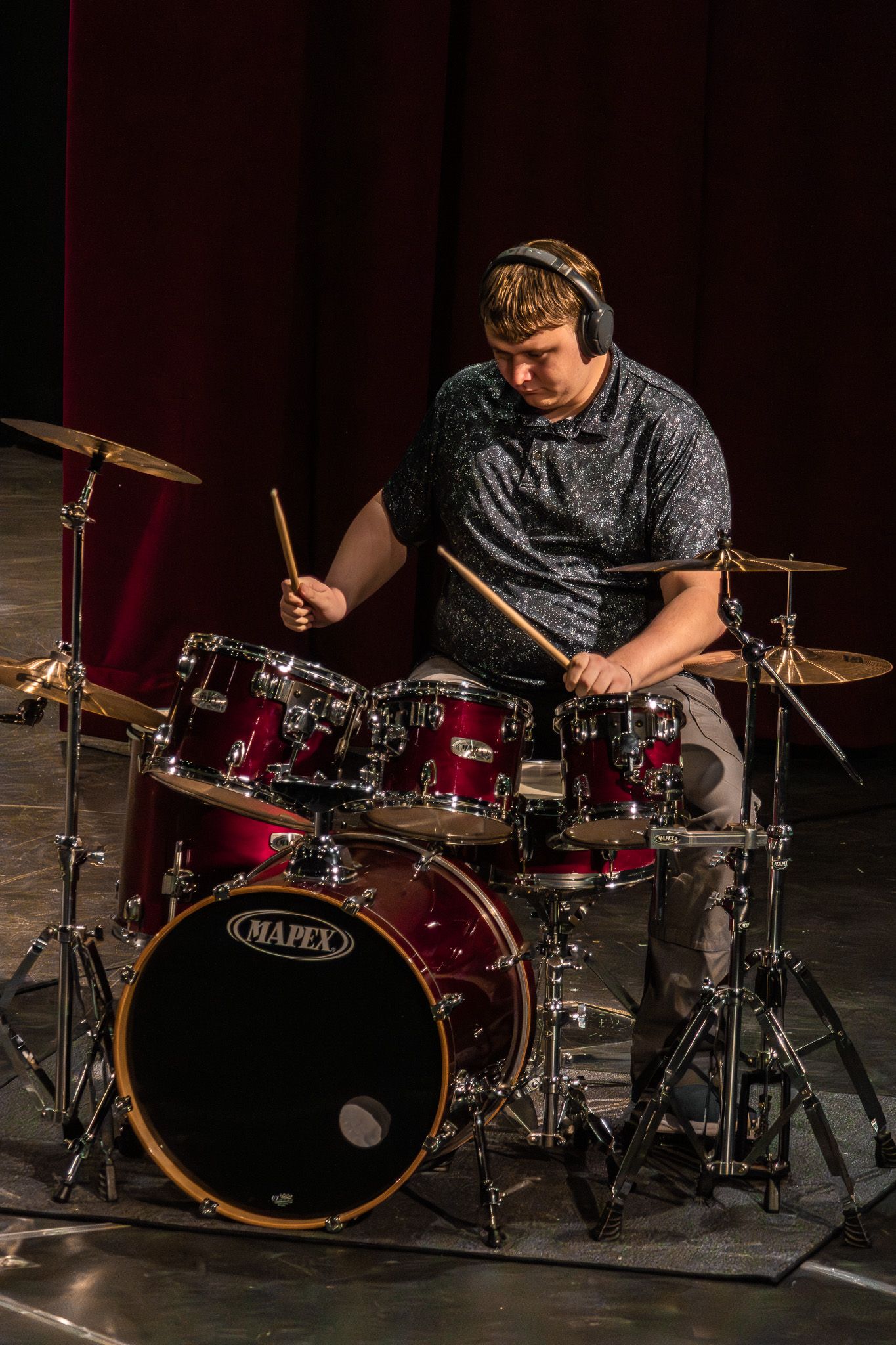 Young male drummer playing red Mapex drum kit on stage wearing headphones at live performance.