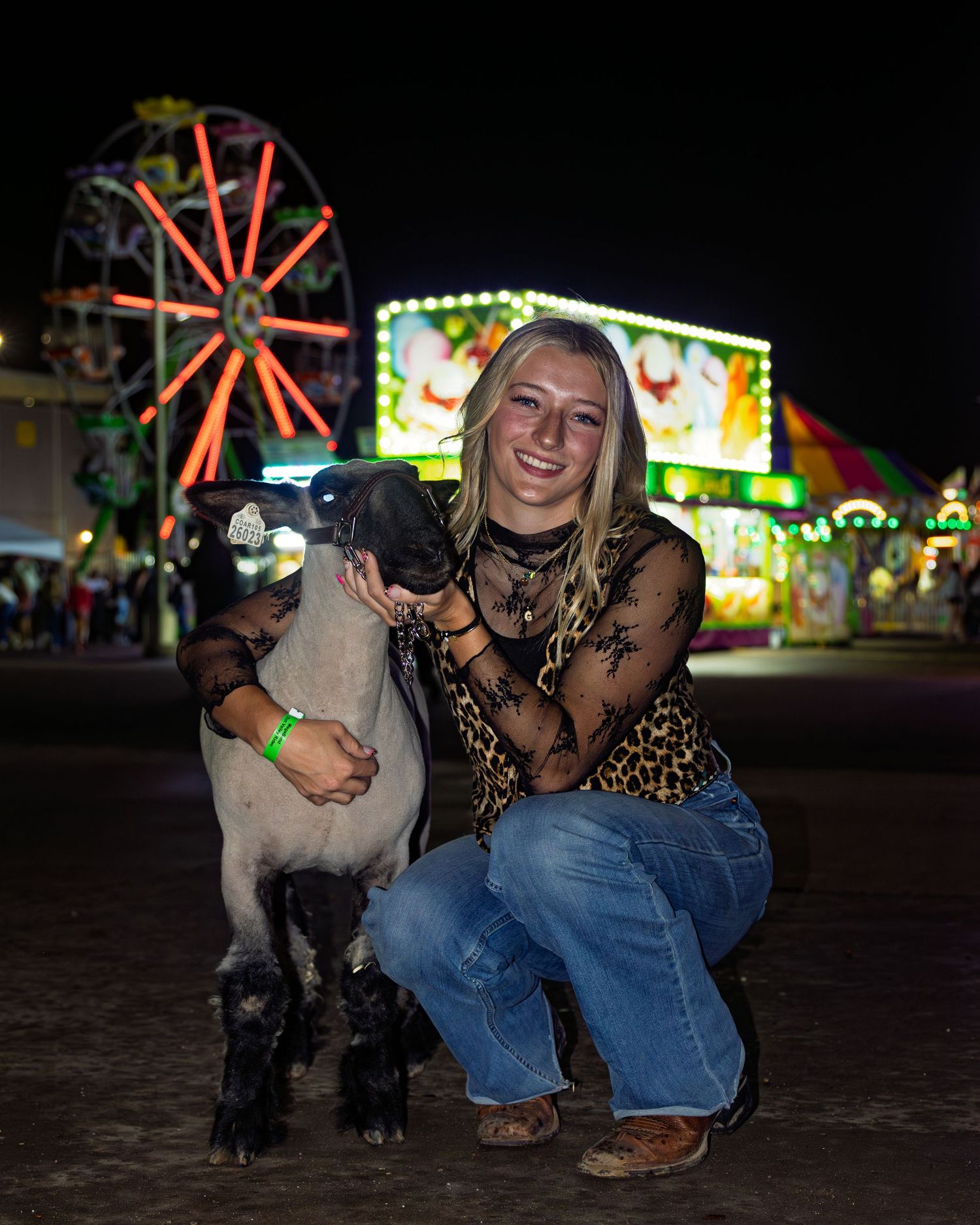 Young woman smiling with a goat at a county fair at night with ferris wheel lights in background.