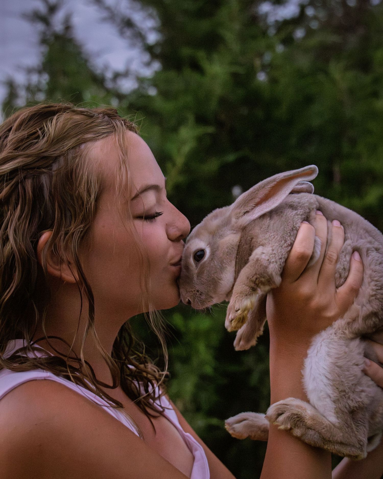 Young woman kissing a large gray rabbit held up in her hands outdoors near green trees.