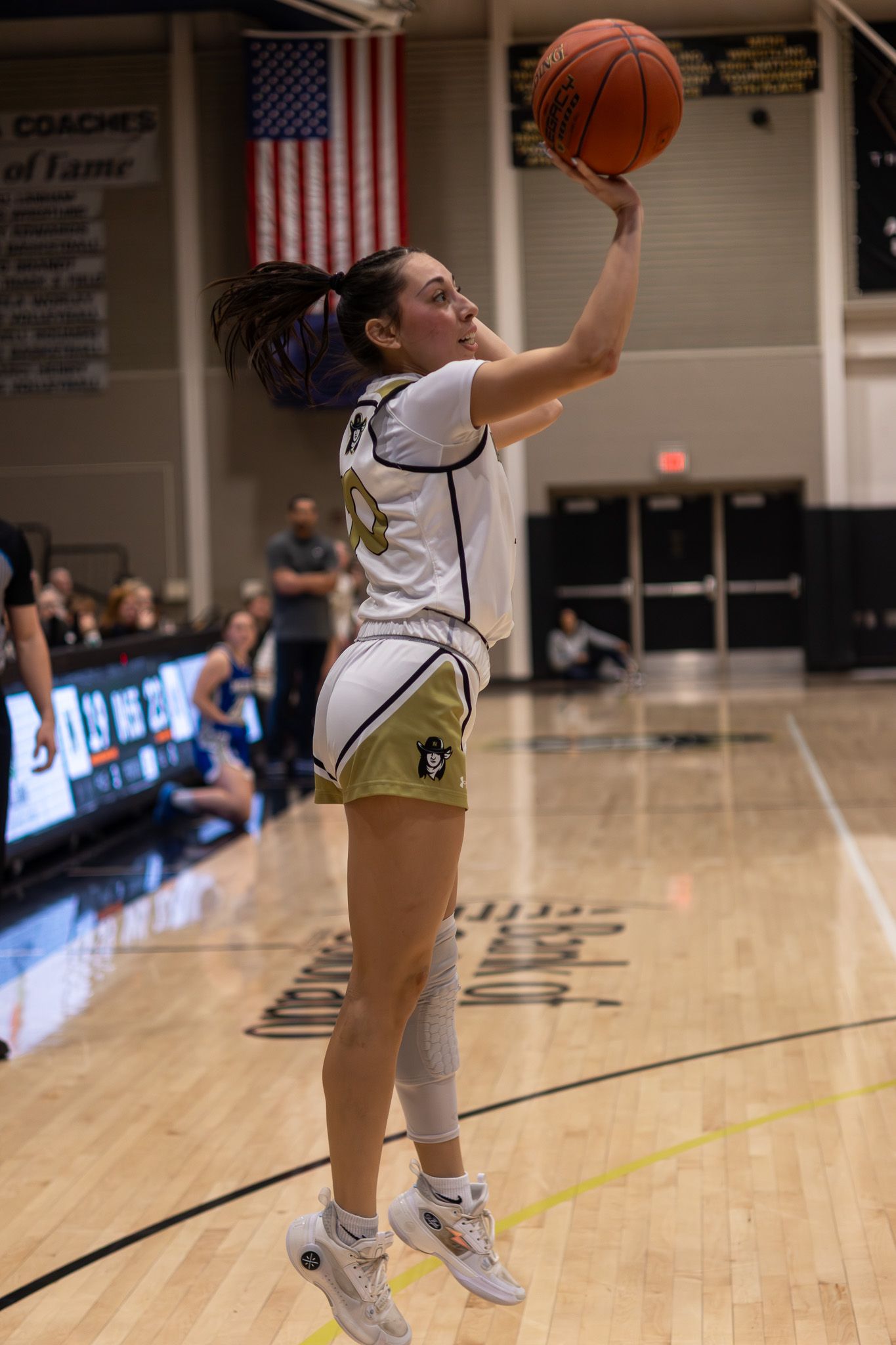 Female basketball player shoots a jump shot during a college basketball game in a white and gold uniform.