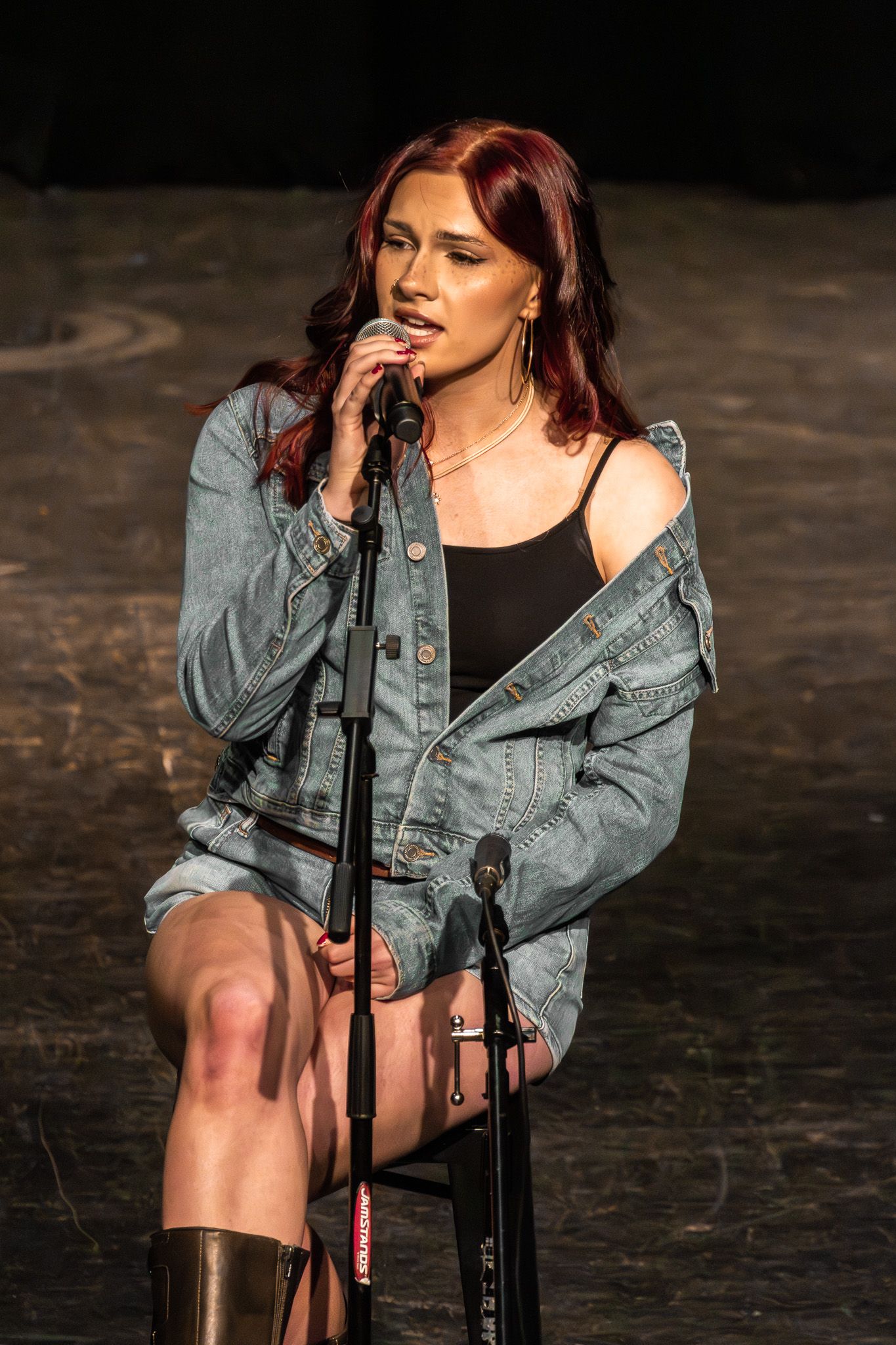 Red-haired female singer performing on stage seated at microphone in denim jacket and black bodysuit.