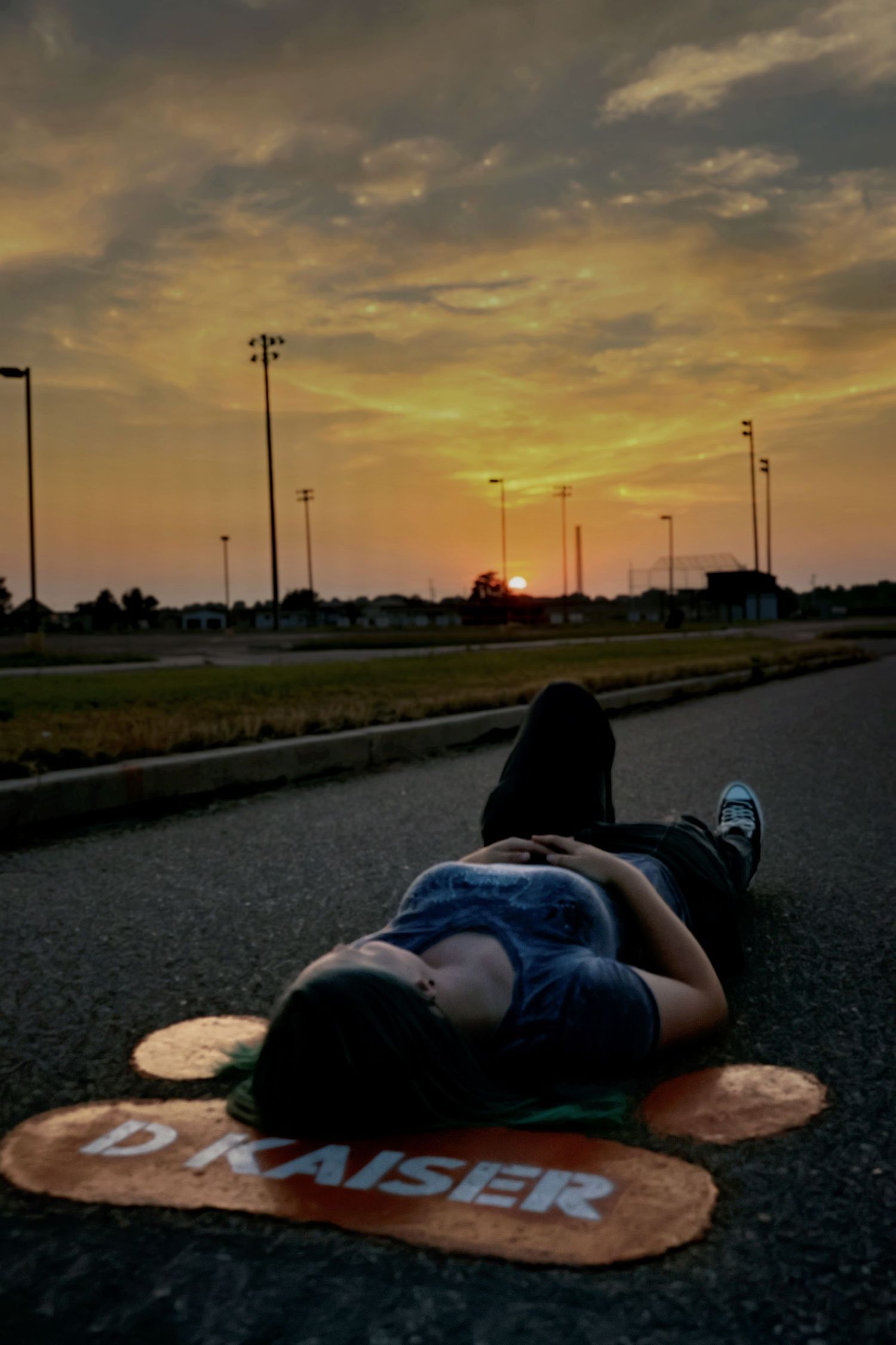 Person lying on a road at sunset near a D Kaiser logo, dramatic golden sky overhead.