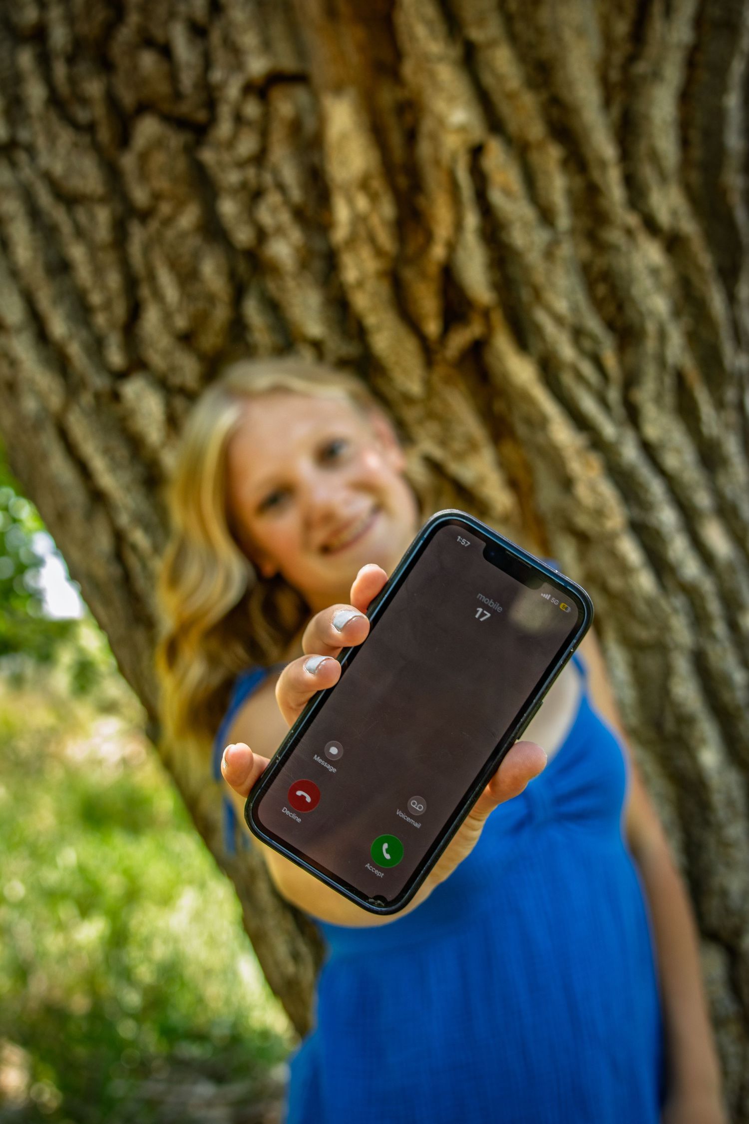 Smiling blonde teen girl holding iPhone showing incoming call near a tree.