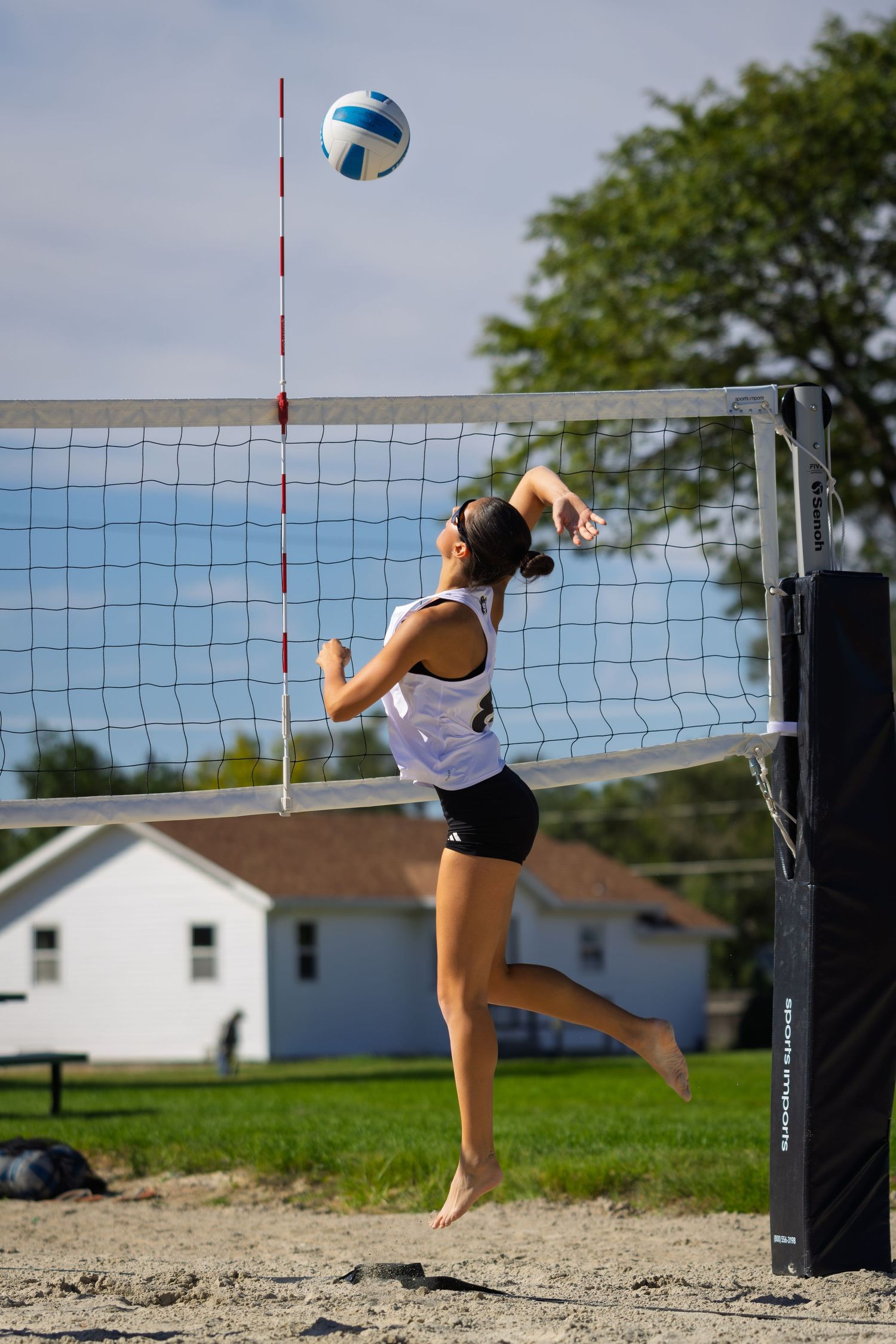 Female volleyball player leaps to spike the ball over the net on a sunny outdoor sand court.