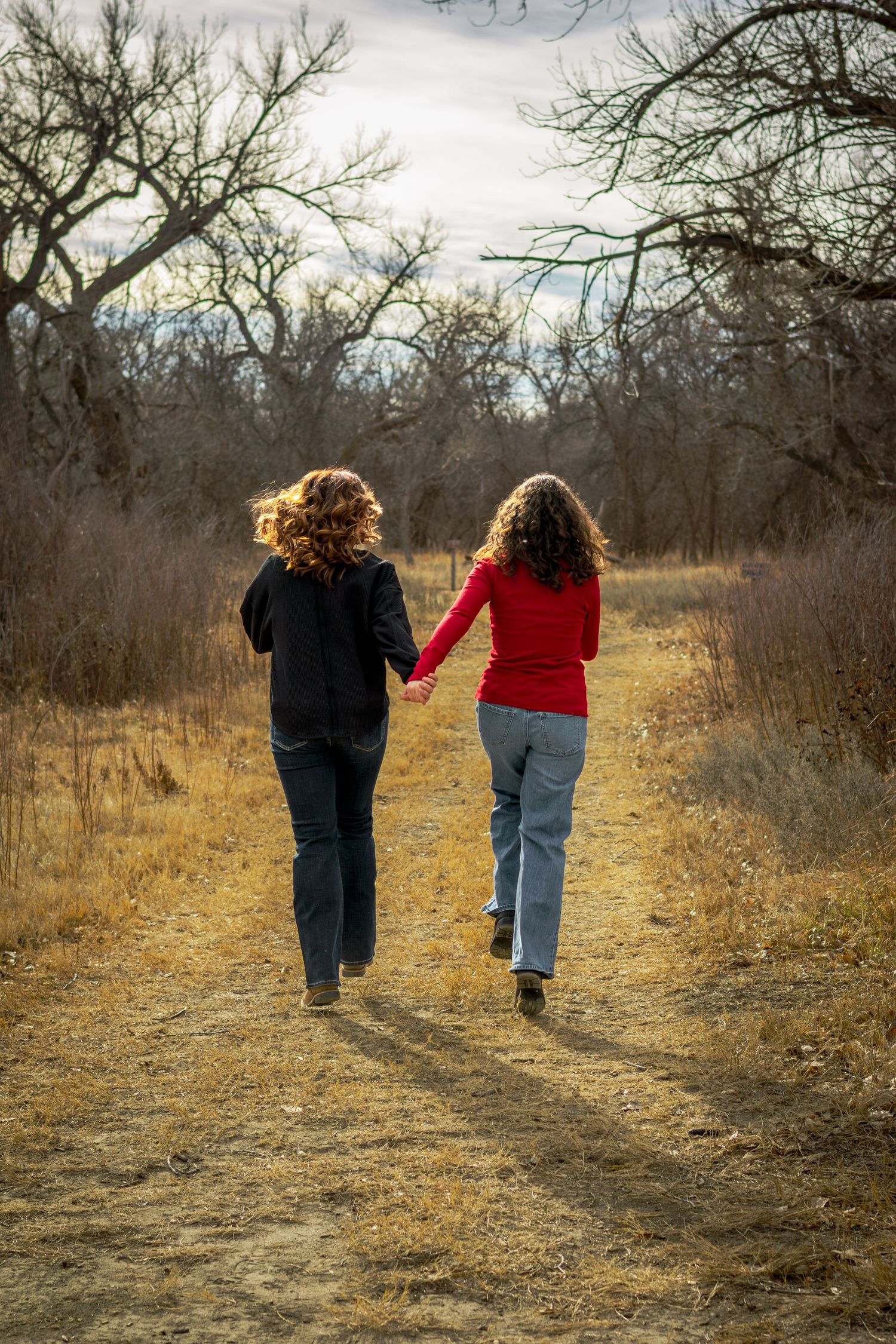 Two curly-haired women holding hands walk away down a dirt path through bare autumn trees.