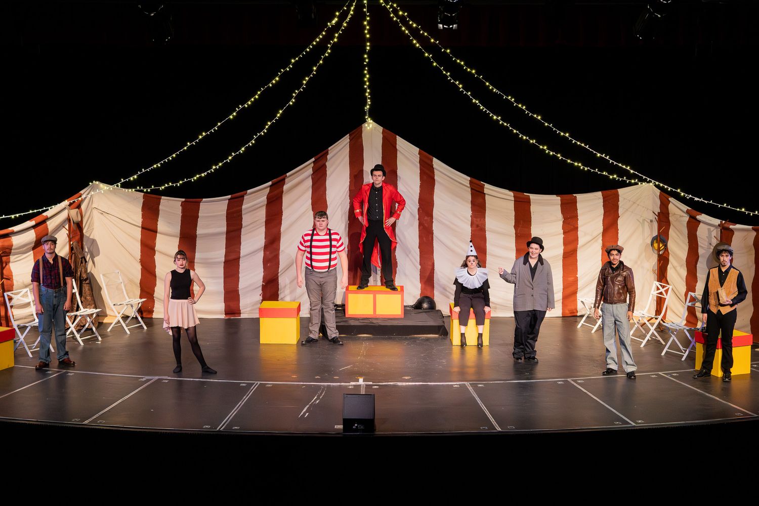 Theater cast performs on stage in front of a red and white striped circus tent backdrop.