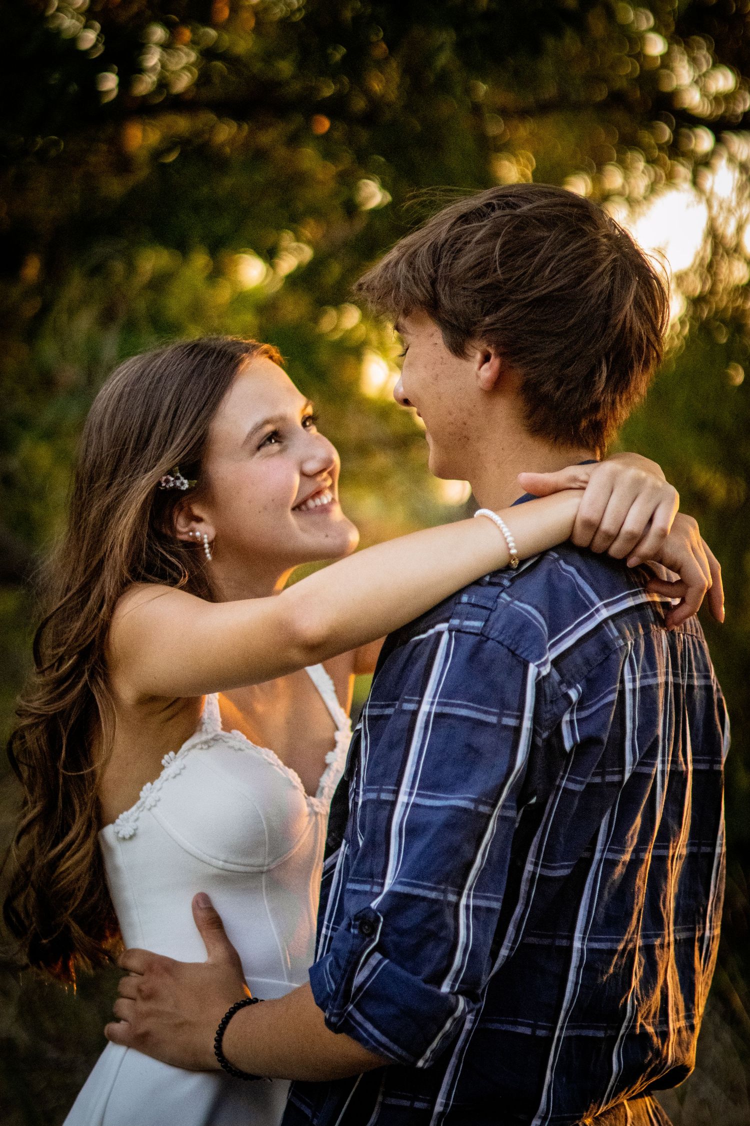 Young couple embracing and smiling at each other in warm golden sunset light among green trees.