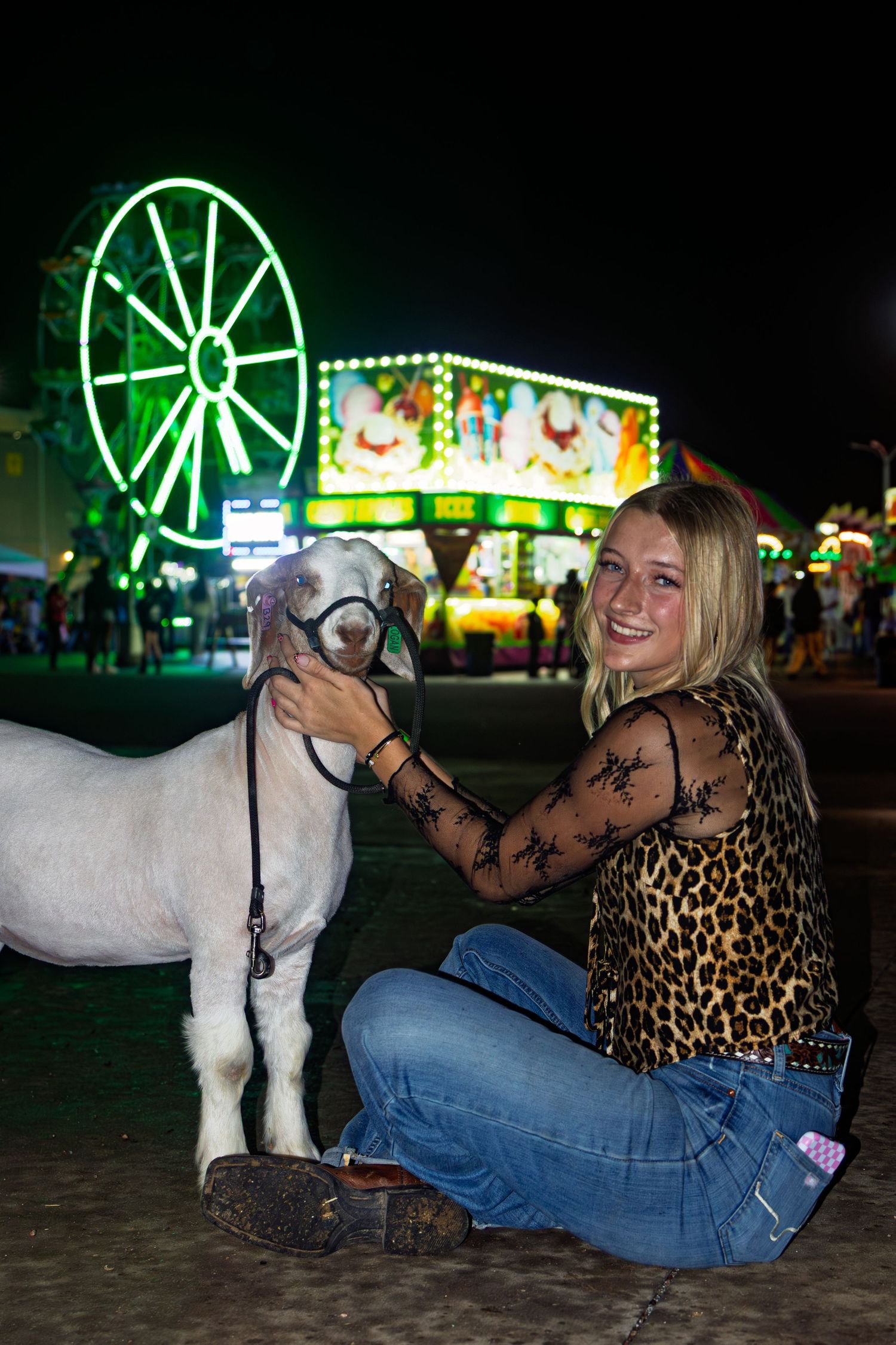 Blonde woman in leopard print top poses with white show goat at illuminated county fair at night.