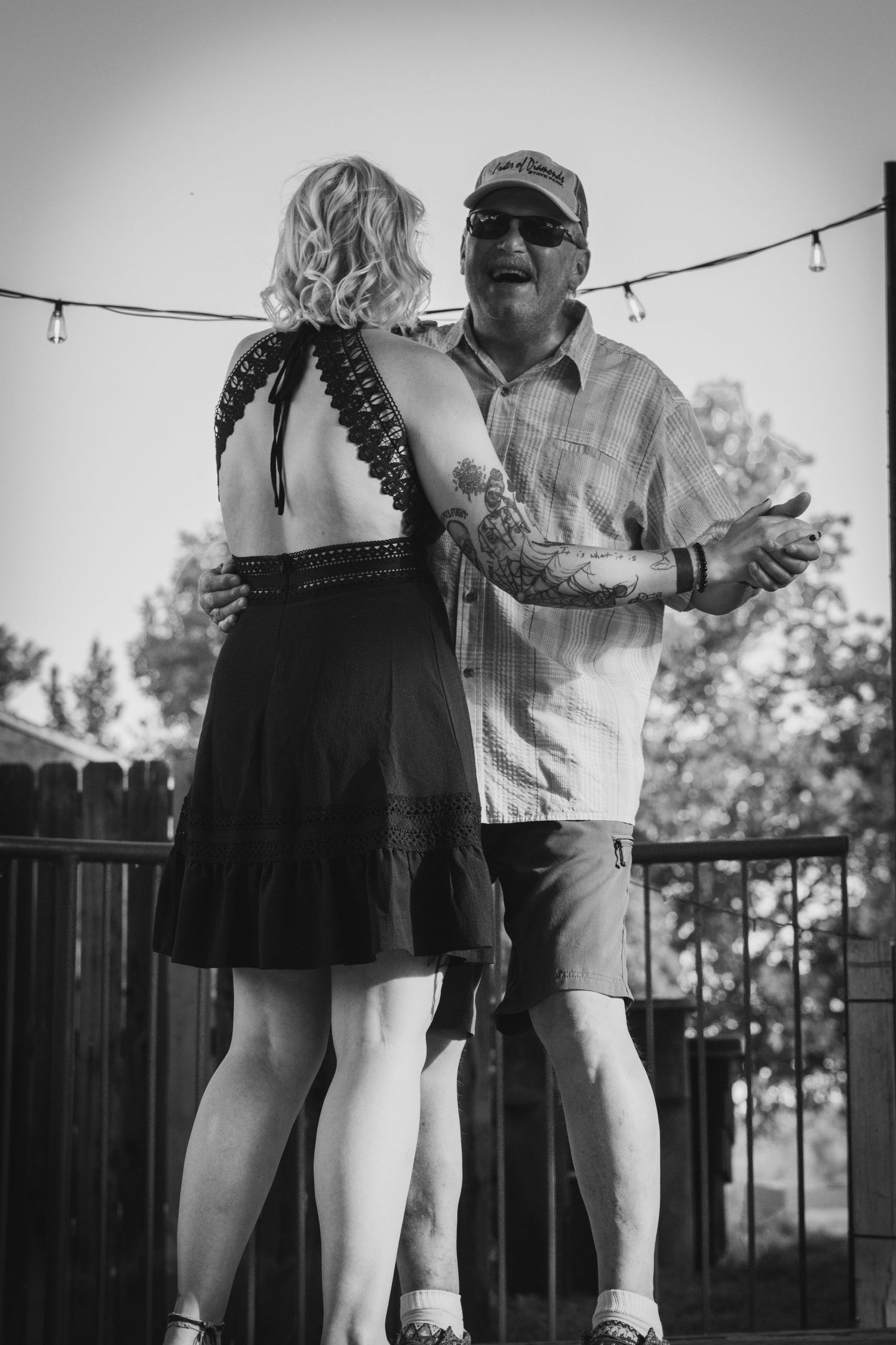 Black and white photo of a couple slow dancing outdoors at a backyard party with string lights overhead.