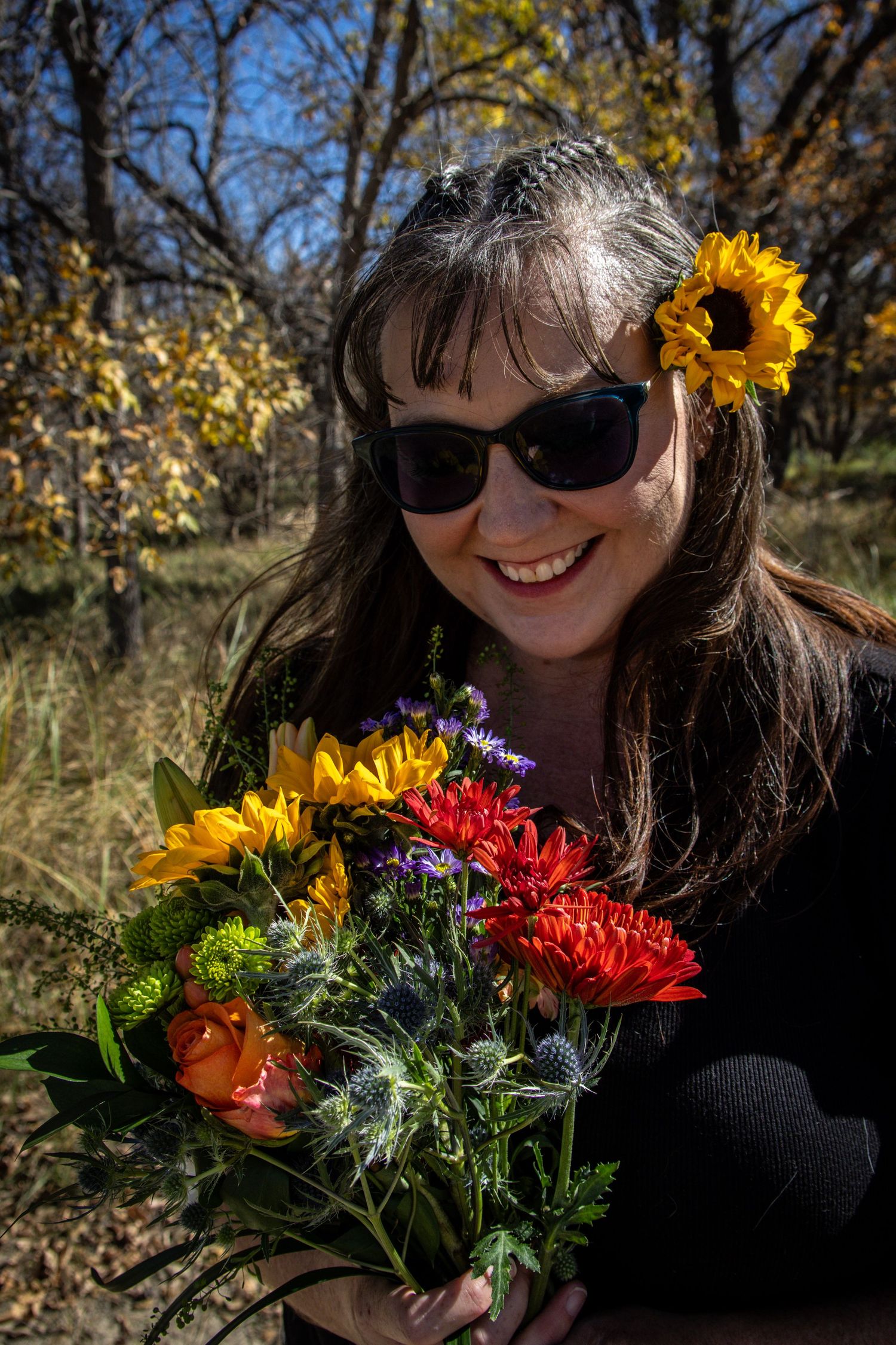 Smiling woman wearing sunglasses holding a vibrant bouquet of sunflowers and mixed flowers outdoors.