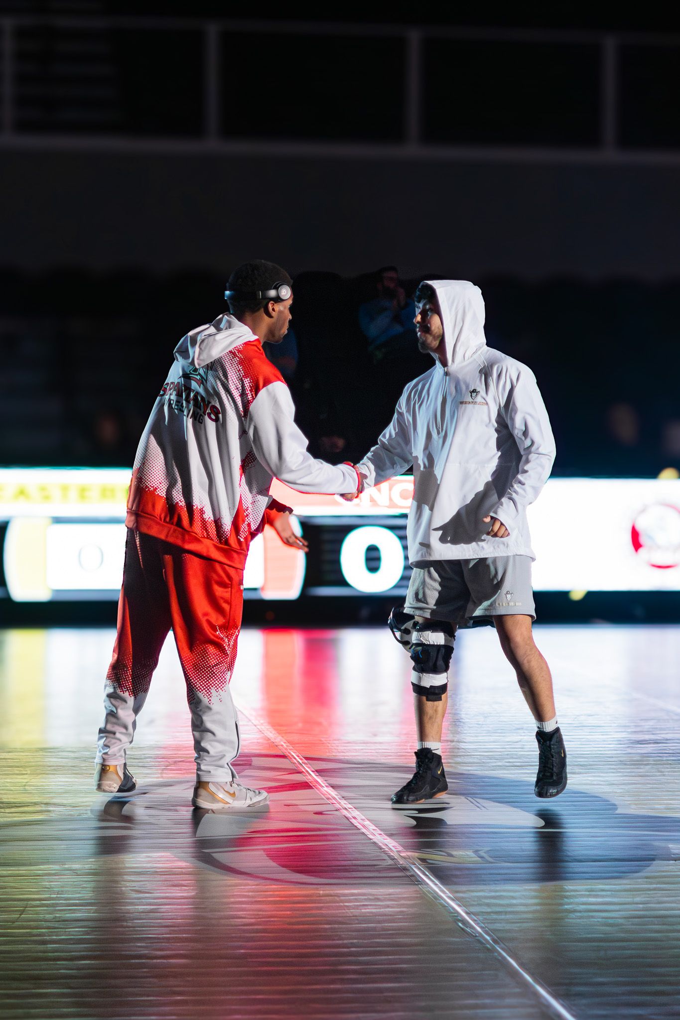 Two athletes in colorful warm-up gear shake hands on an indoor court before a wrestling match.