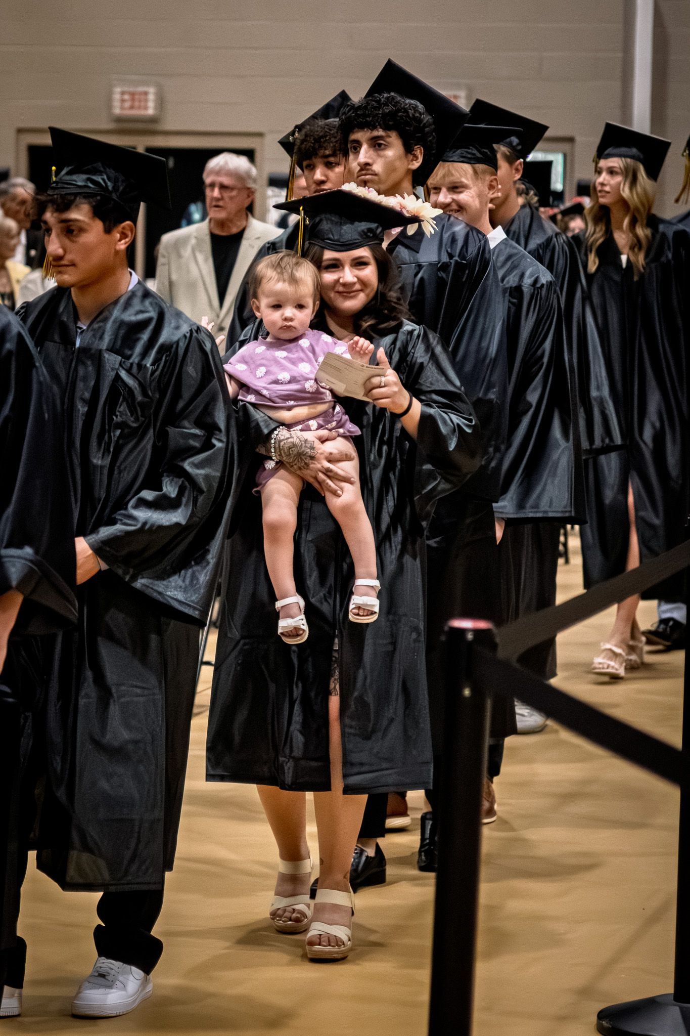 Smiling graduate in black cap and gown holding baby during graduation ceremony procession.