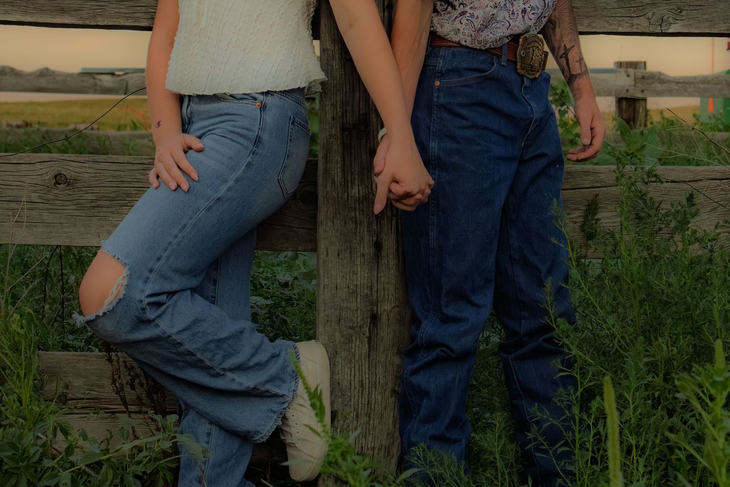 Close-up of couple holding hands while leaning against a rustic wooden fence post in a green field.