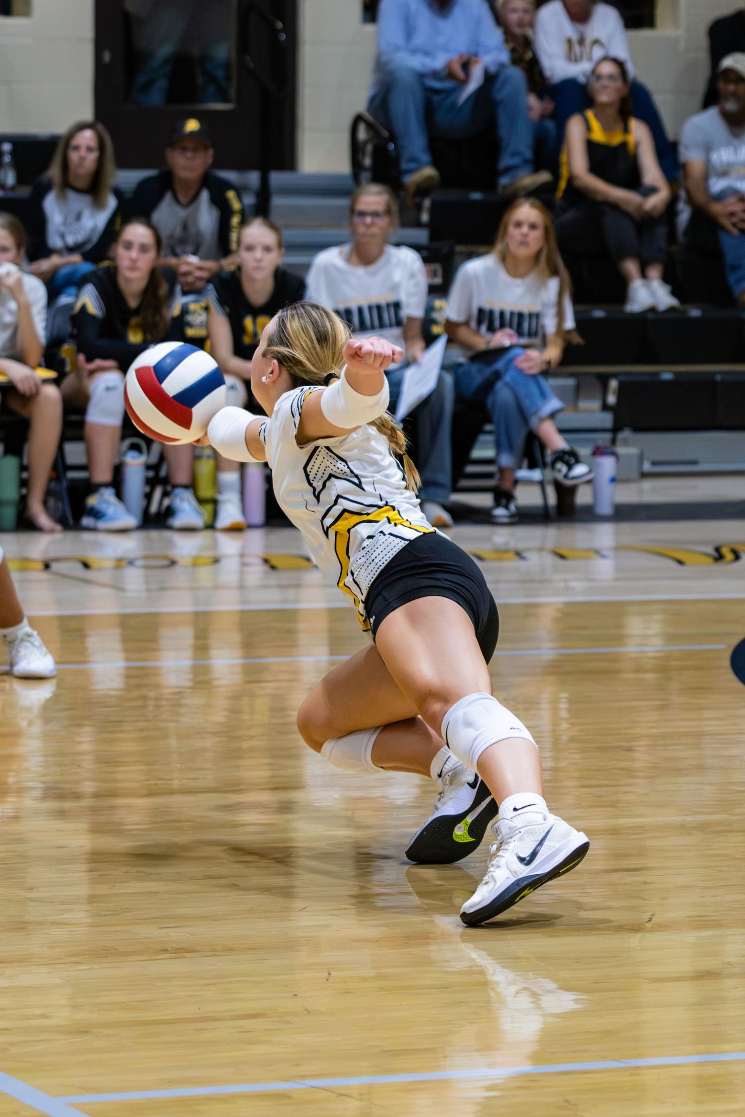 Female volleyball player in black and gold uniform diving low to dig the ball during a high school match.