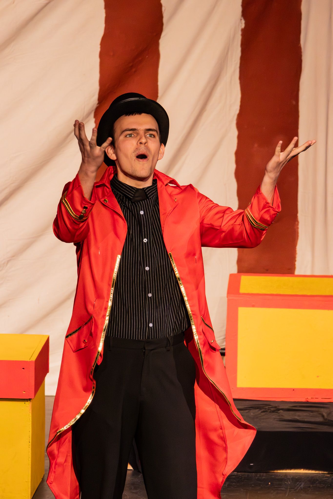 Male performer in red ringmaster coat and black top hat gestures dramatically on stage.