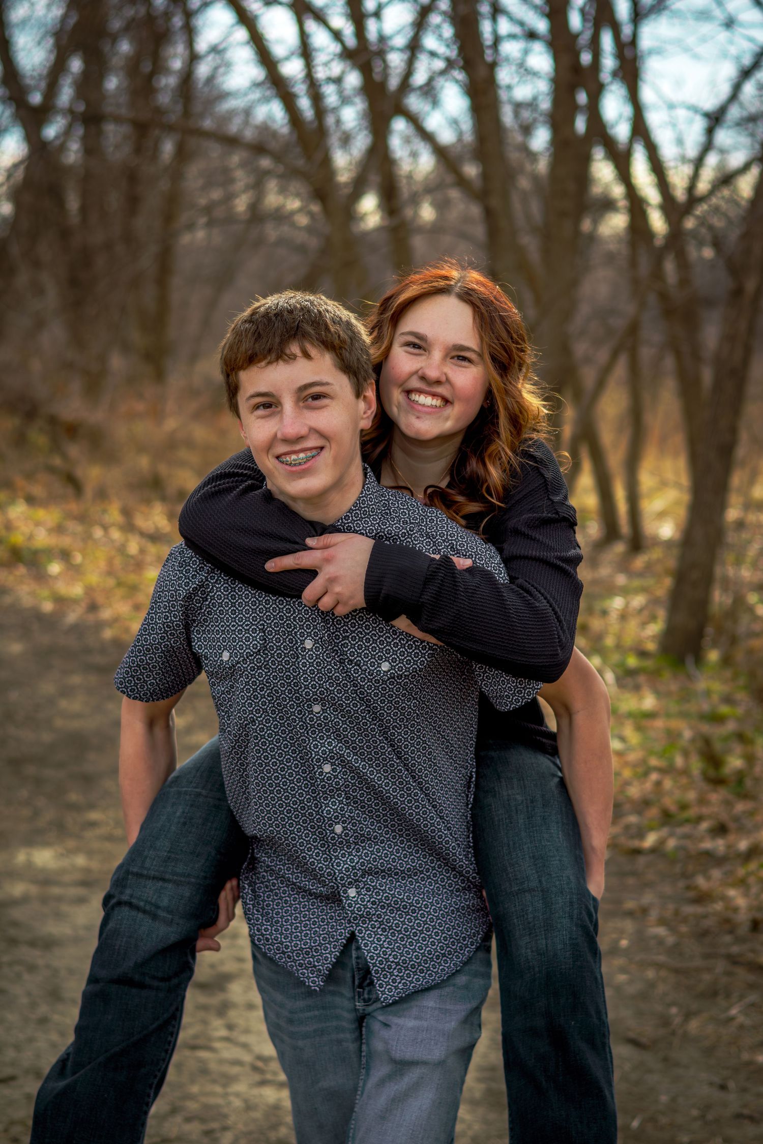 Happy teenage boy giving a smiling red-haired girl a piggyback ride on a wooded trail.