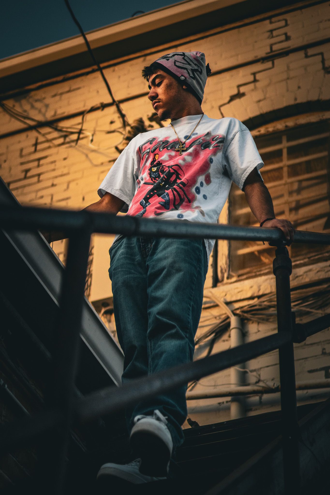 A young man in a graphic tee and beanie leans on a metal railing on outdoor stairs at dusk.