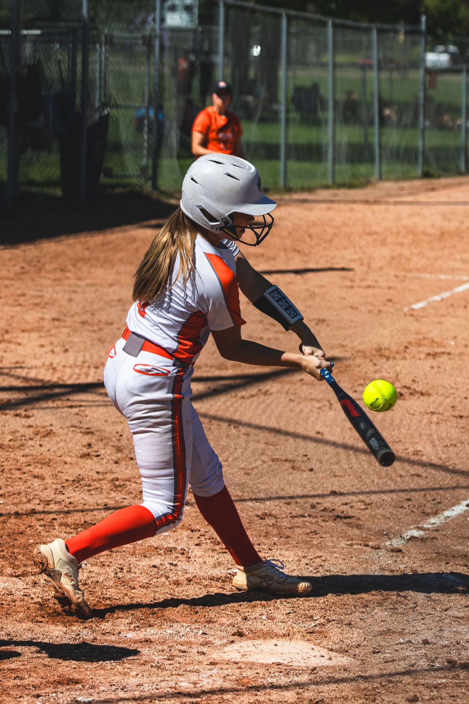 Female softball player in white and orange uniform swinging at a yellow pitch near home plate.