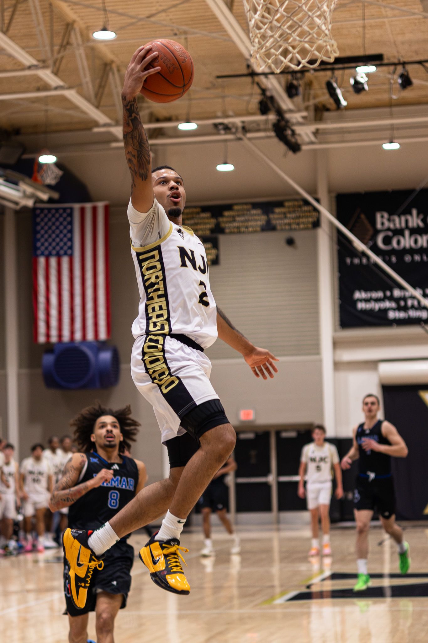 NJC Plainsmen player #2 goes up for a layup in a gold and white uniform during a college basketball game.